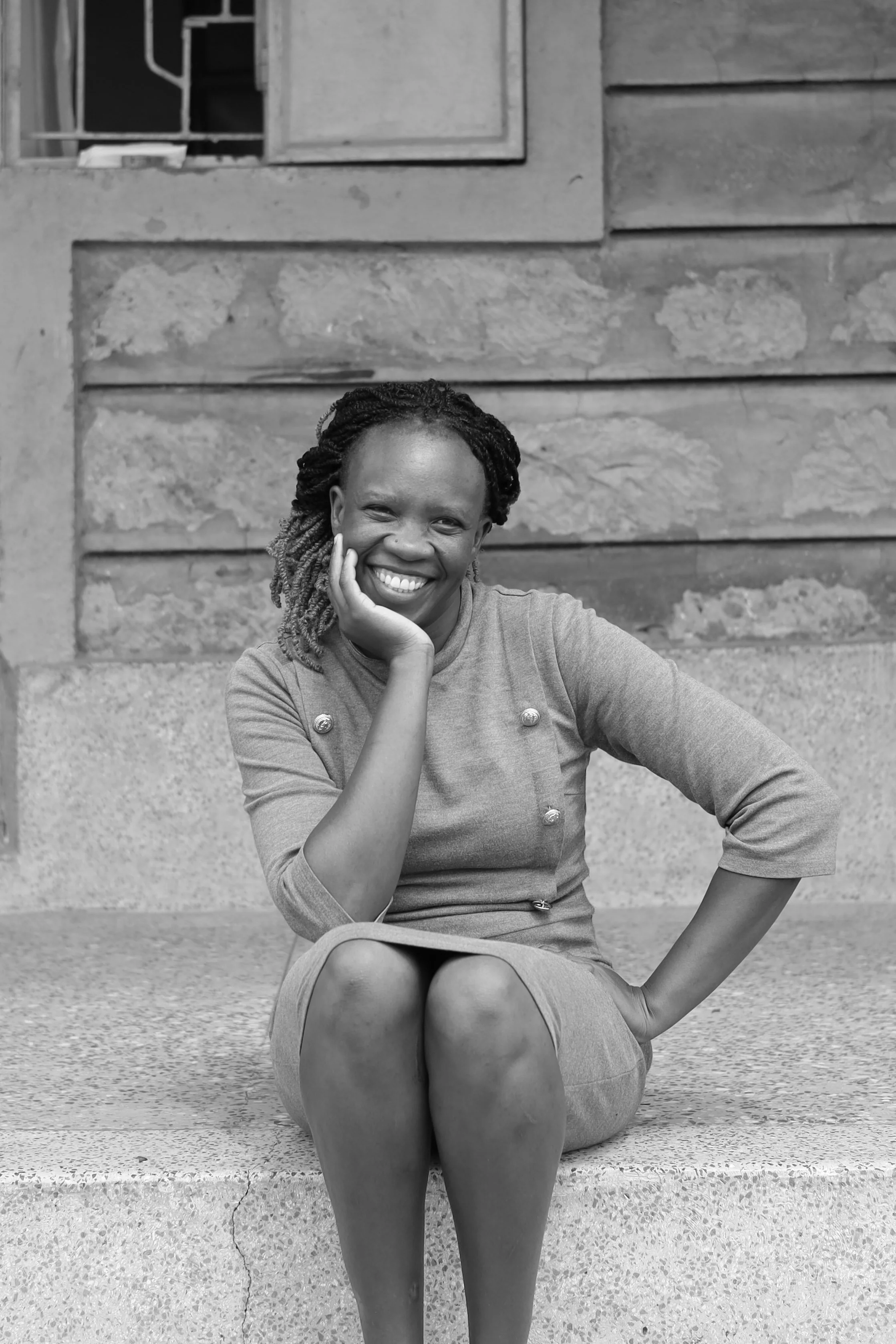 A woman with long braided hair sitting on a concrete step, smiling and resting her chin on her hand, in front of a textured stone and wooden building.