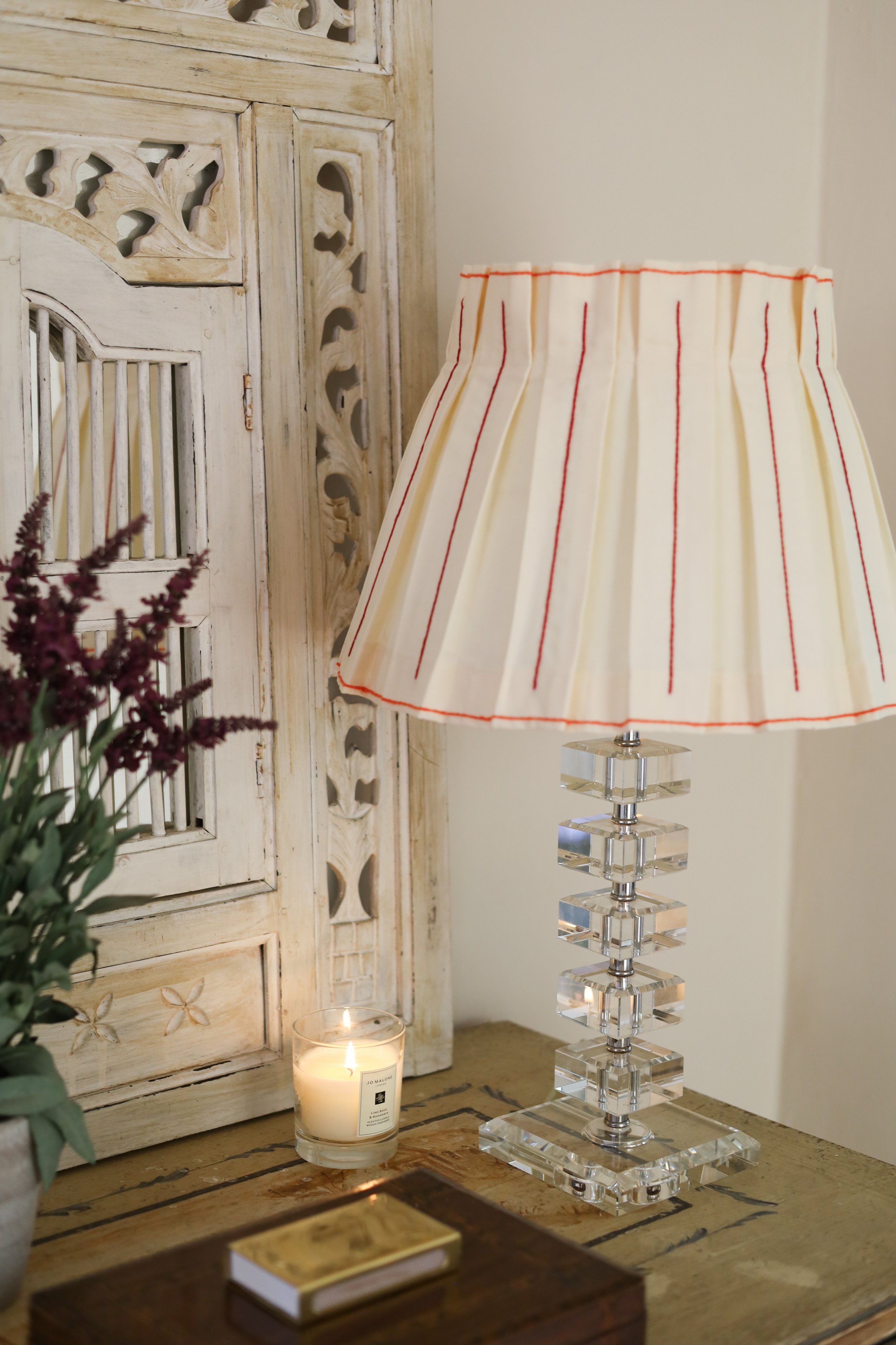 A decorative table lamp with a crystal base and a pleated fabric shade on a wooden table, next to a lit candle in a glass container, a small potted plant, and a closed book in a cozy interior setting.