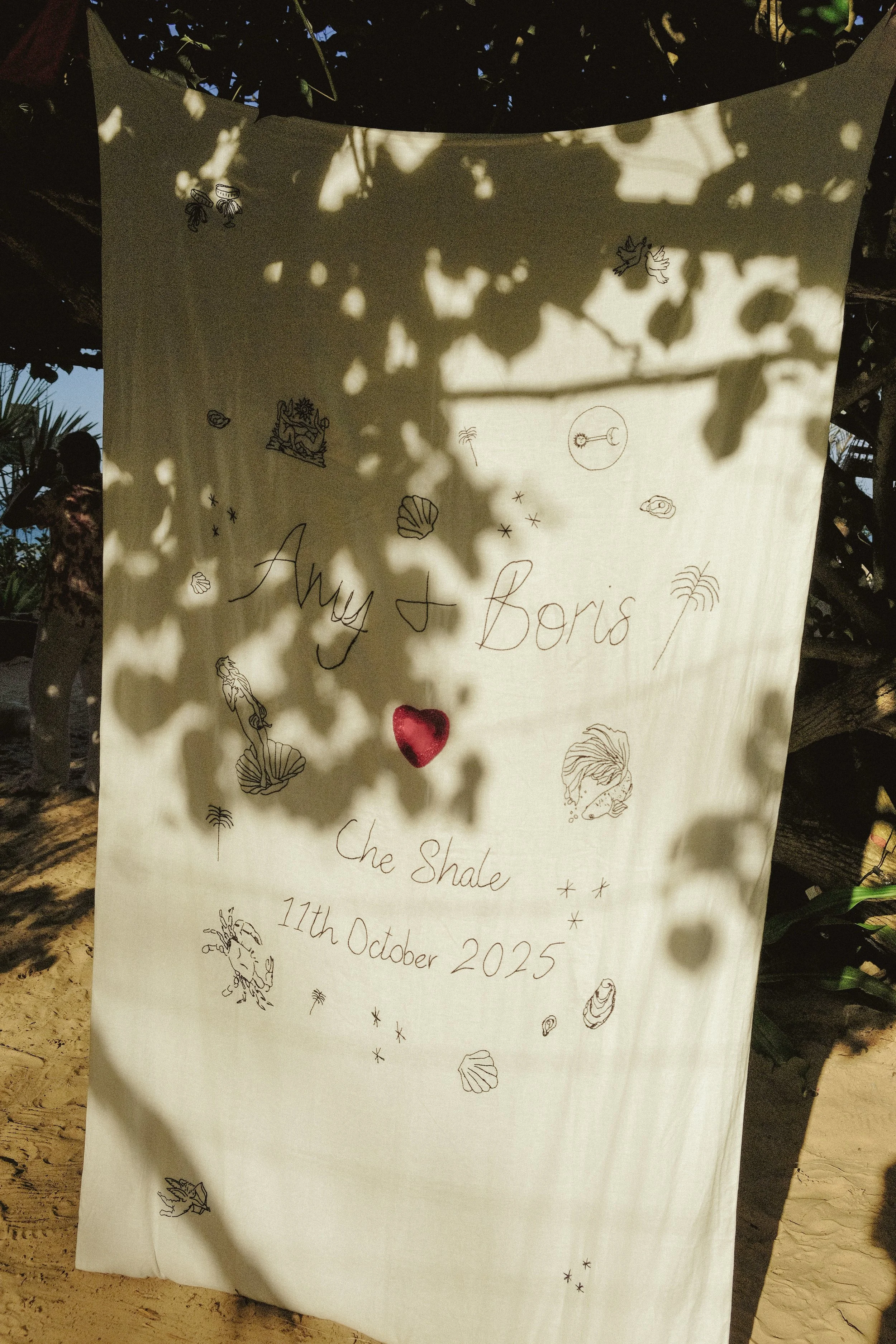 A decorative sign with handwritten text that reads 'Amy & Boris' and 'che Shale 11th October 2025', surrounded by small drawings of sea shells, a crab, a mermaid, and ocean-related items, hanging outdoors with shadows of tree leaves cast on it.