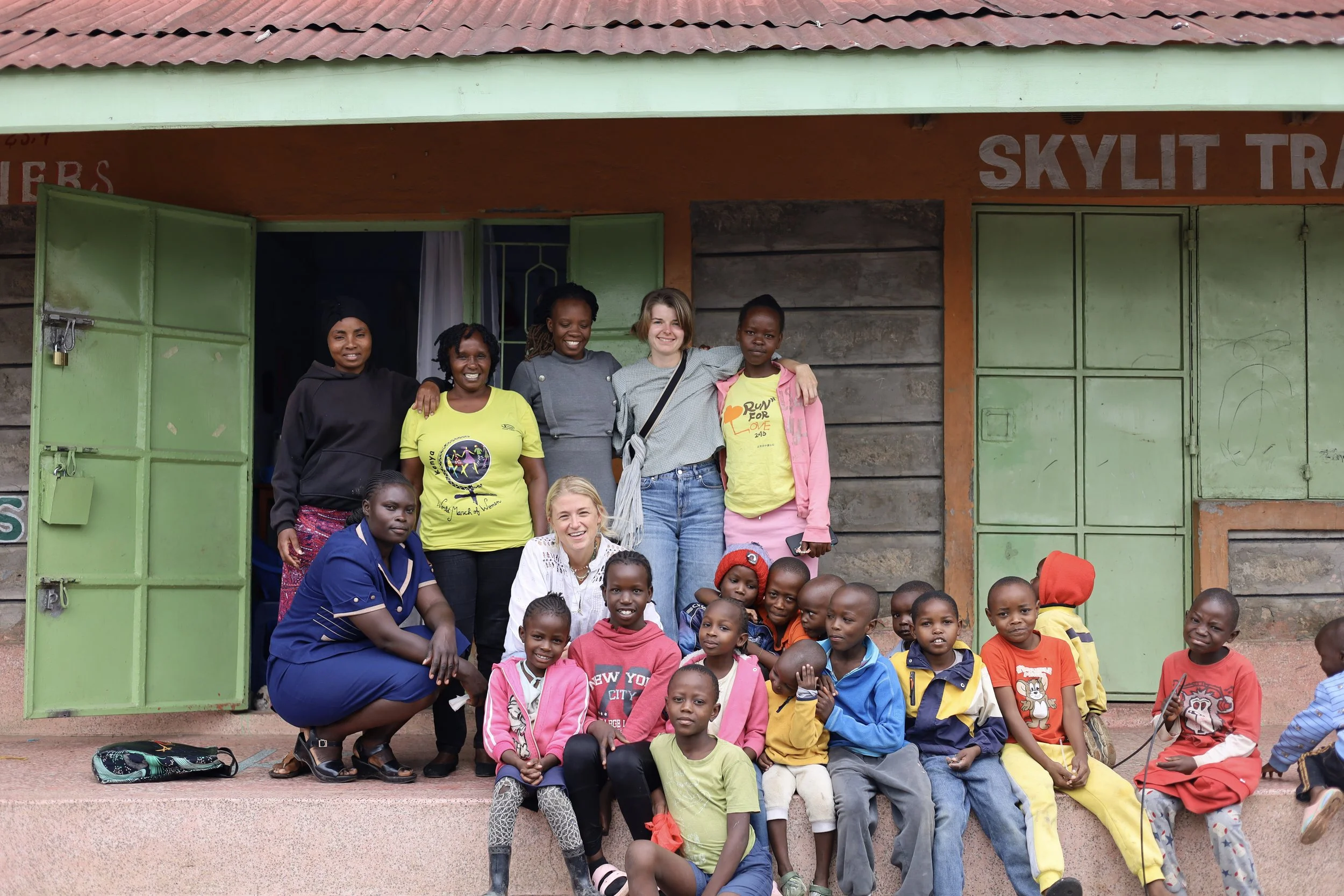 Group of smiling children and women posing outside a building with green doors and a red roof.