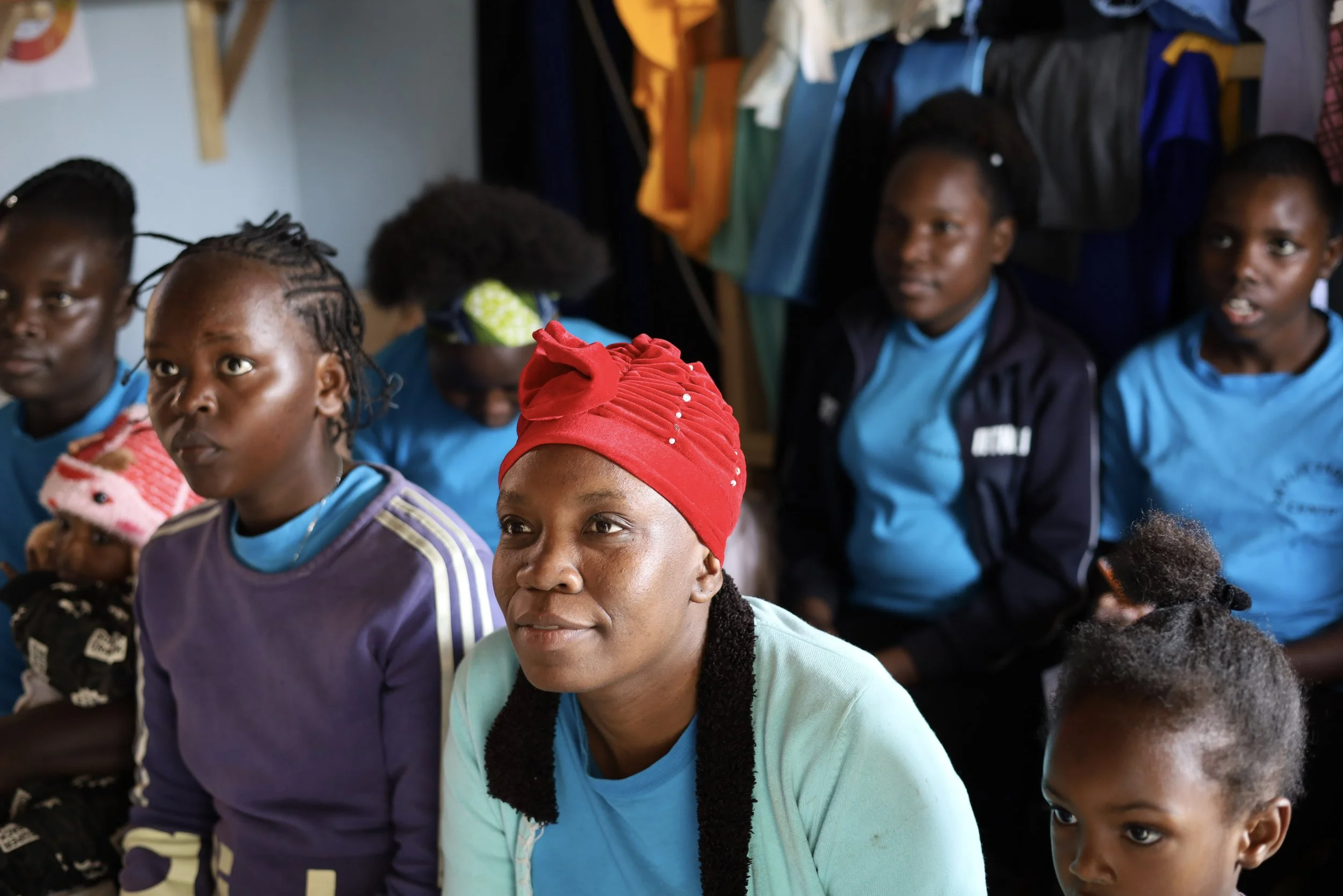 A group of women and girls sitting indoors, listening attentively during a gathering or event. One woman in the foreground wears a red headscarf, and others are dressed in blue and purple clothing.