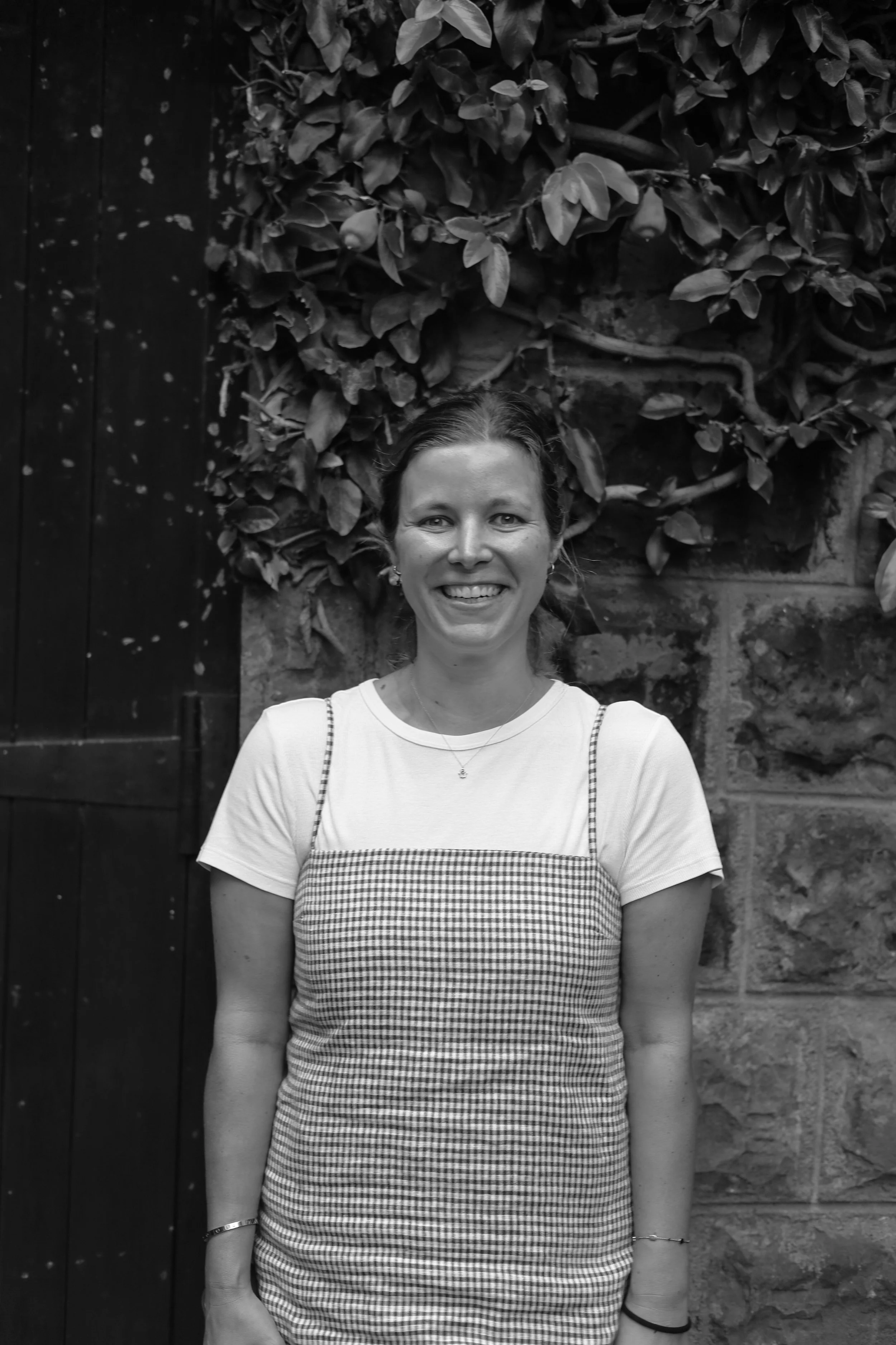 A woman smiling outdoors in front of a brick wall with a leafy vine growing on it.