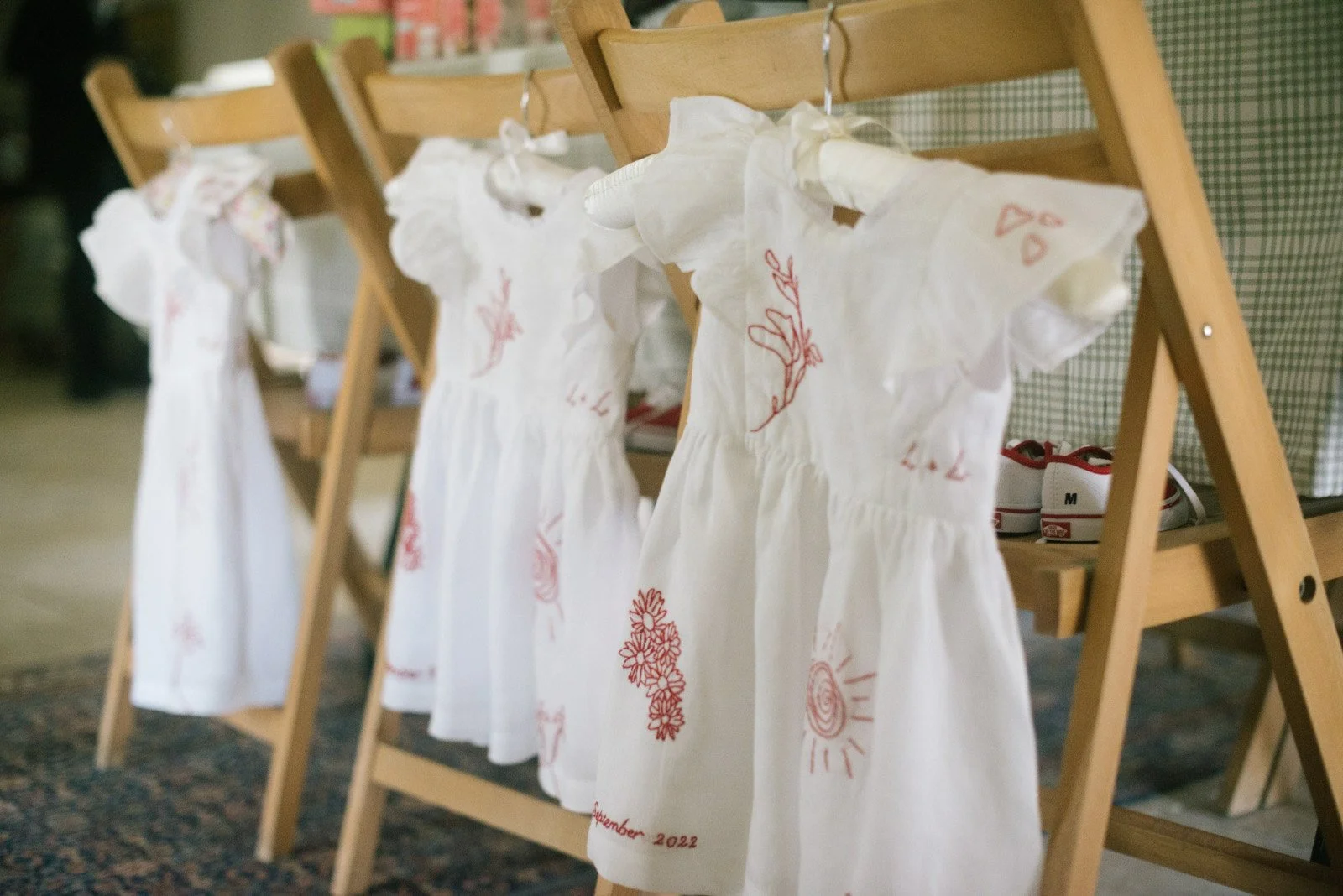 Four white dresses hanging on a wooden rack, each with red embroidered designs and text, in a room with a floral rug and shelves with shoes and other items in the background.