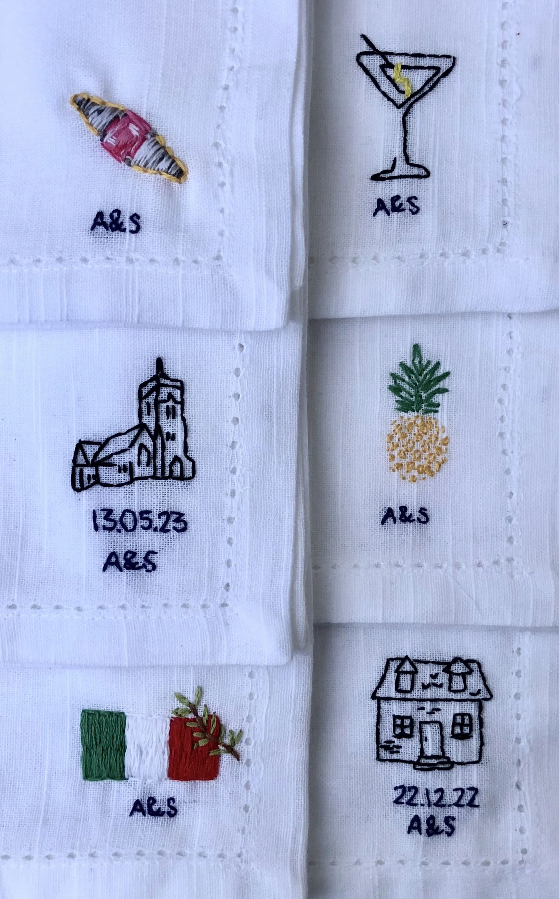 Six white fabric patches with embroidered images and text: a seashell with 'A&S', a cocktail glass with 'A&S', a church with date '13.05.23' and 'A&S', a pineapple with 'A&S', an Italian flag with 'A&S', and a house with date '22.12.22' and 'A&S'.