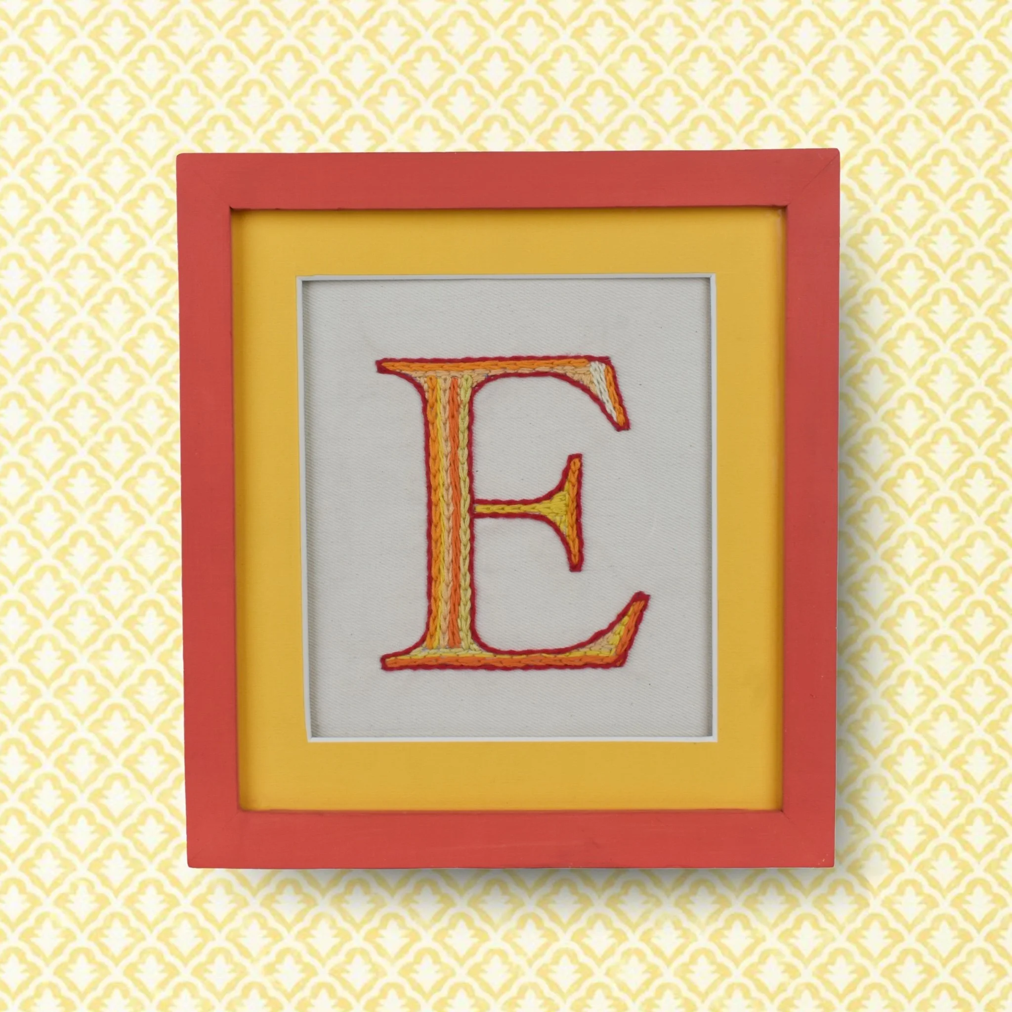 Decorative embroidered letter E framed in a pink and yellow frame, hanging on a yellow and white floral-patterned wall.