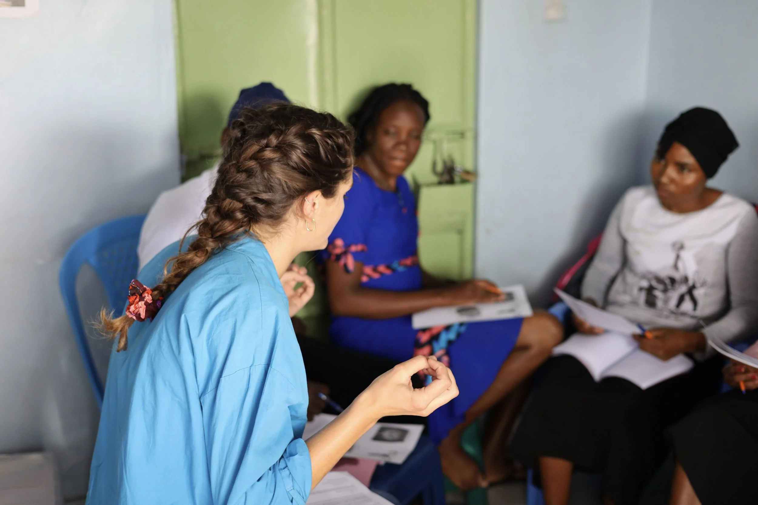 Healthcare professional talking to two women in a small room, one woman wears a black head covering and gray shirt, the other woman wears a blue dress; all are sitting on chairs with documents.