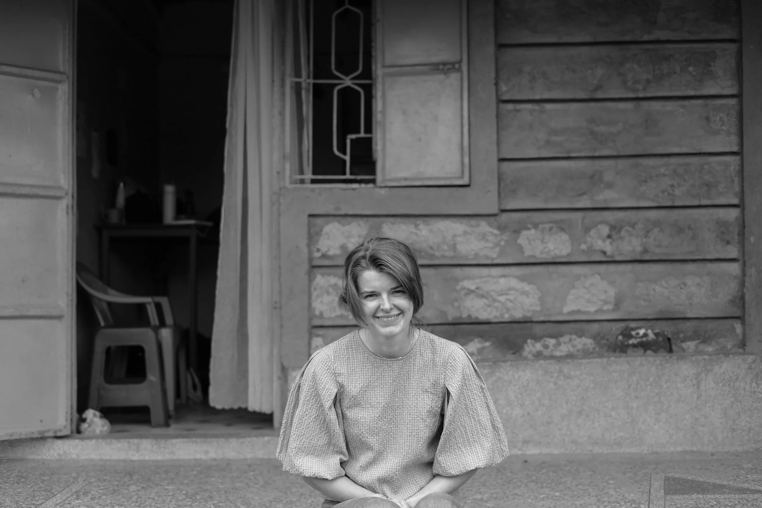 A young woman with short hair waving and smiling, sitting on the ground outside a wooden house, black and white photograph.