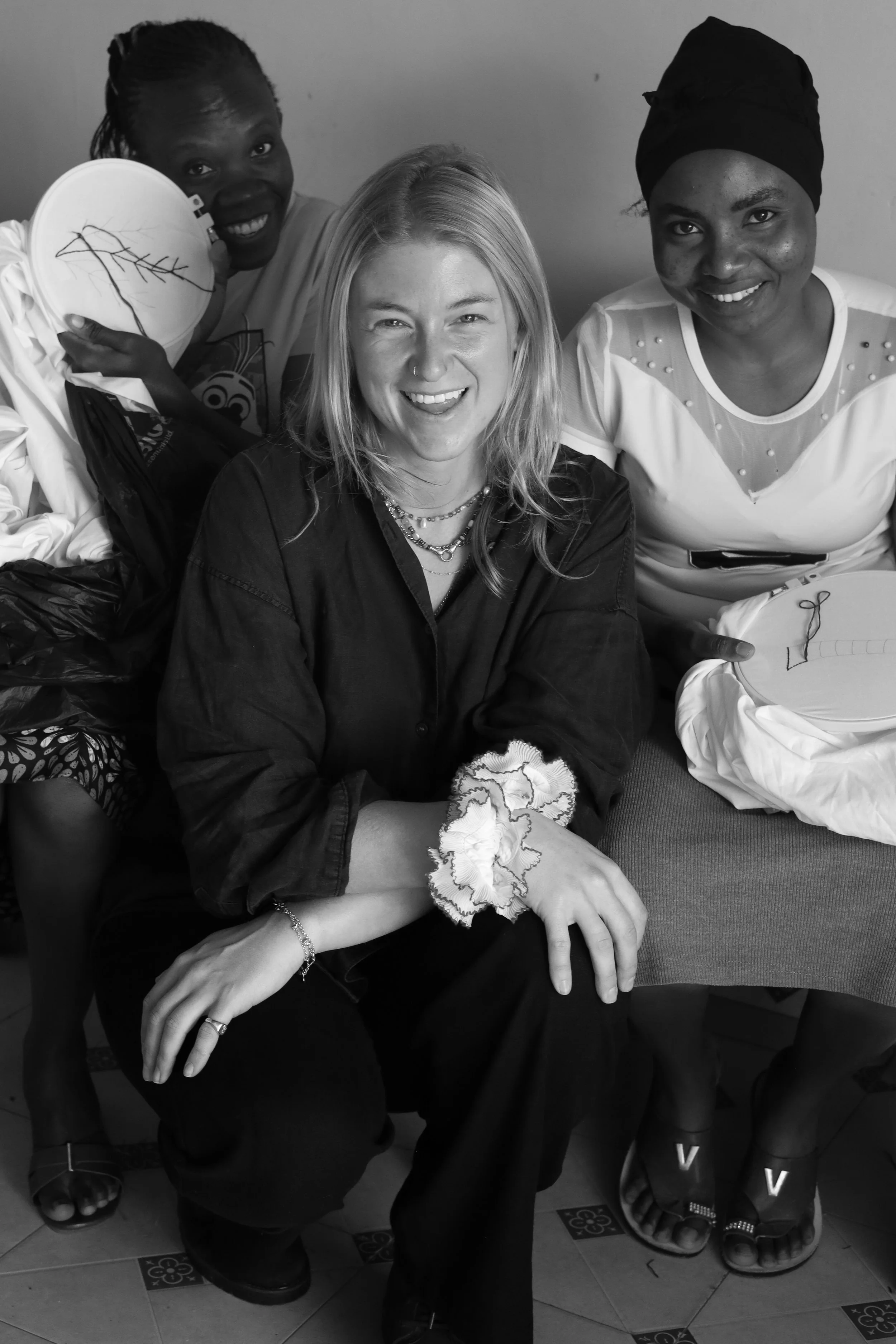 Three women smiling, with one woman sitting in front and two women standing behind her, one holding a pillow with a stitched branch design, in a casual indoor setting.