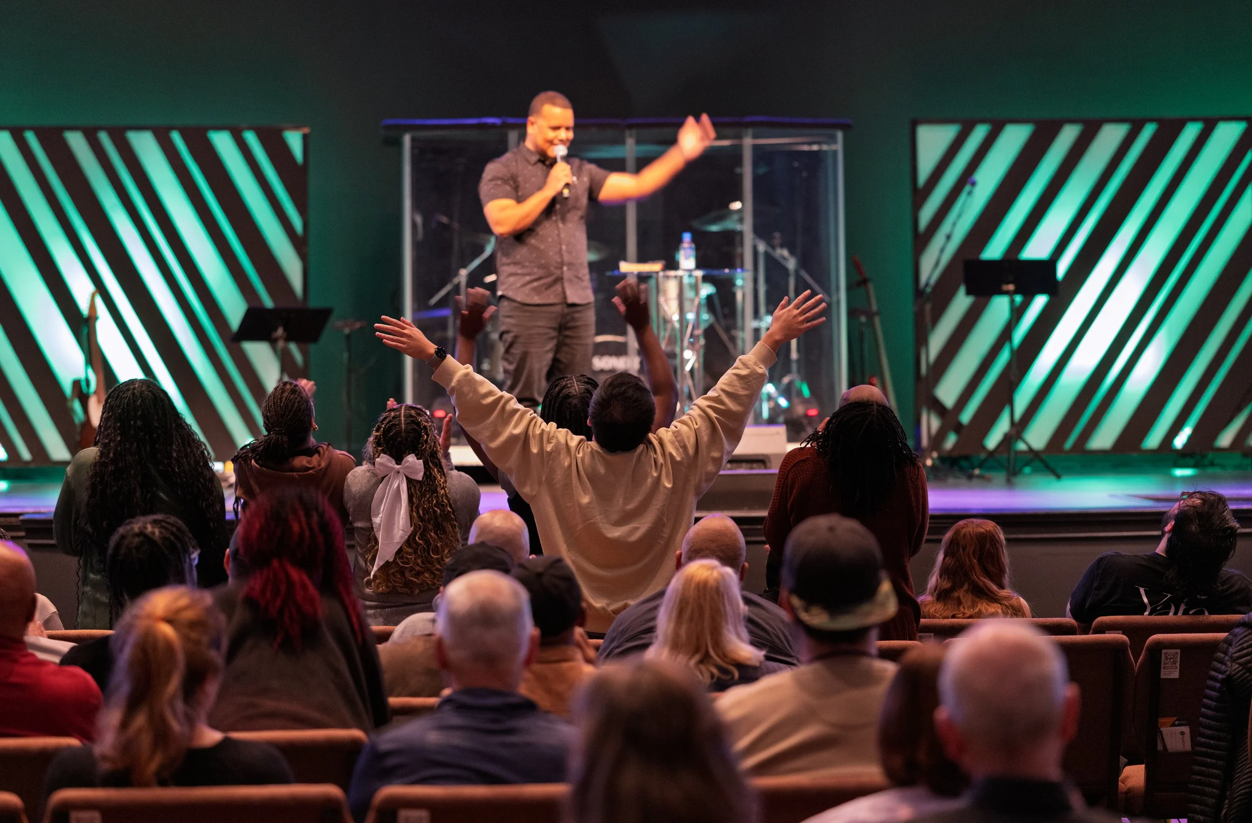 A man on stage leading a religious worship service with raised hands, facing seated congregation in a church or auditorium. The stage has musical instruments and decorative panels with green lighting.