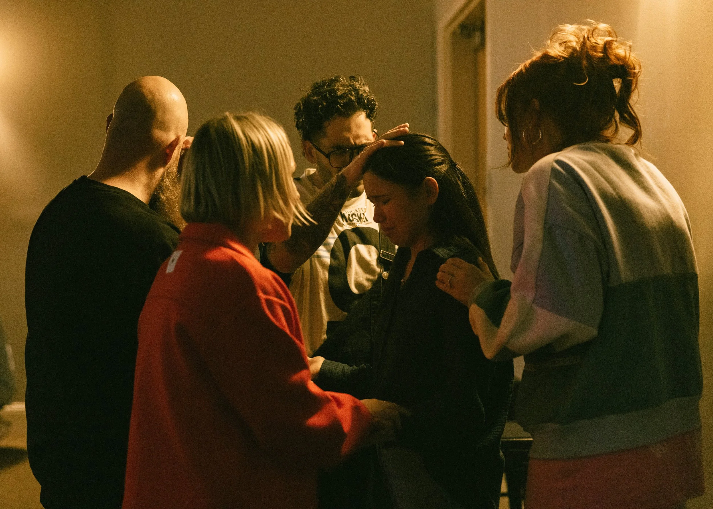 Group of six people gathered closely, with one person placing a hand on another person's head in a moment of prayer or comfort.