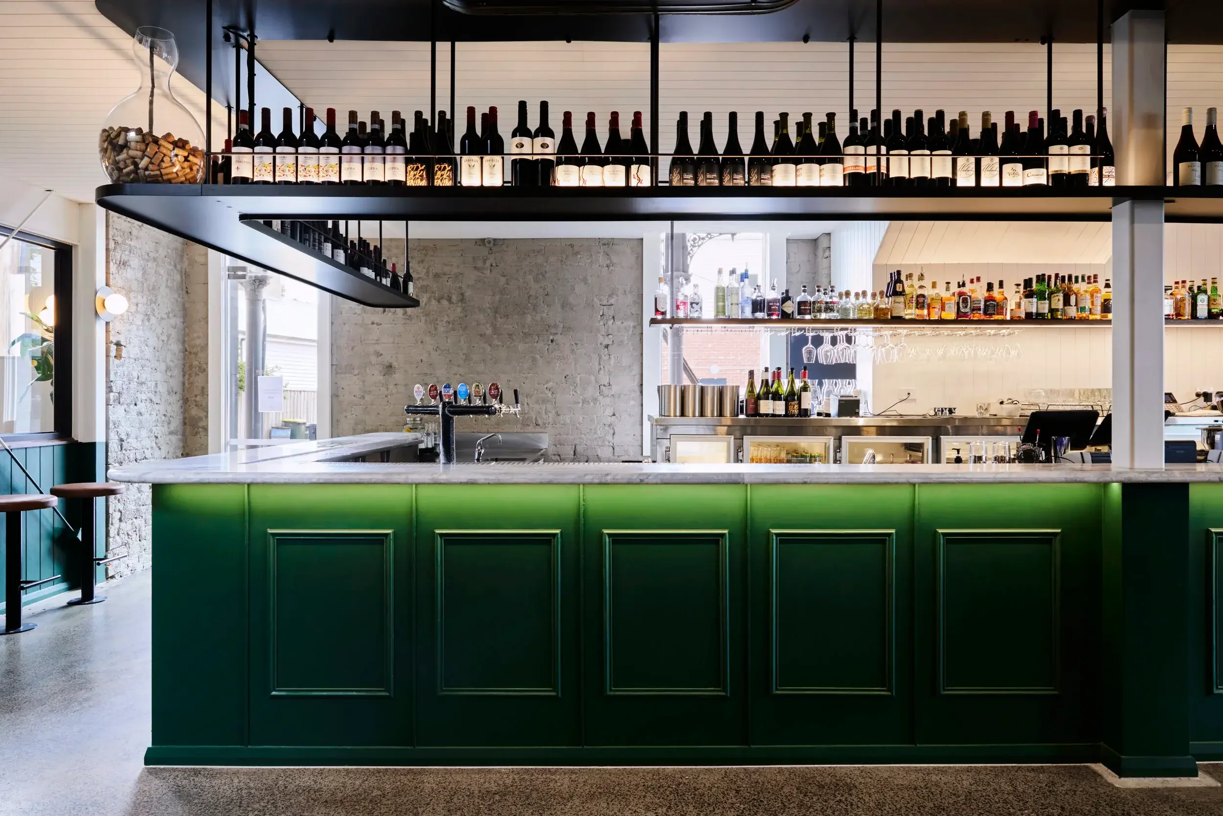Modern bar interior with green paneling, wine bottles on display, and a well-stocked back bar.