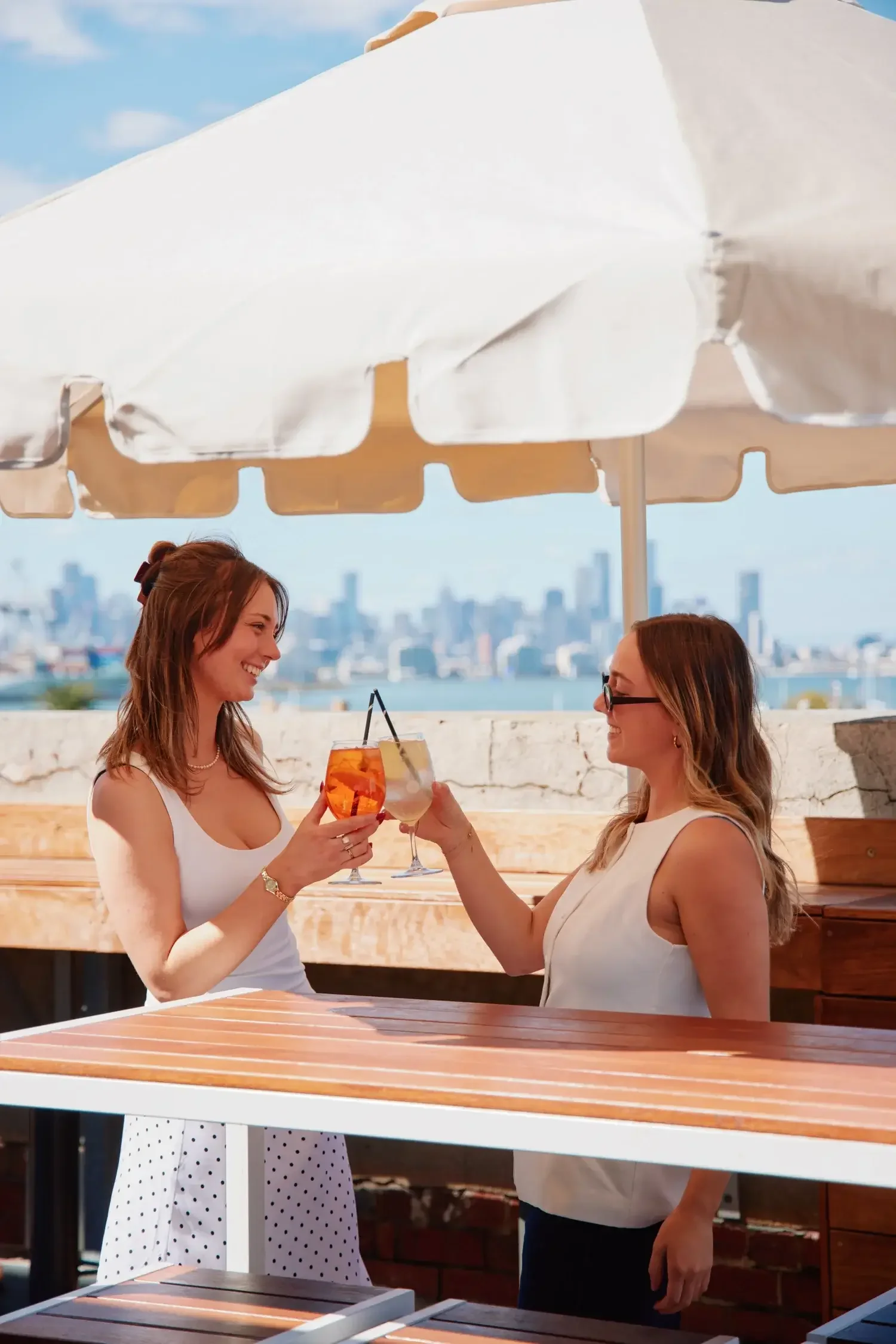 Two women toasting with cocktails under a sun umbrella with a city skyline in the background.