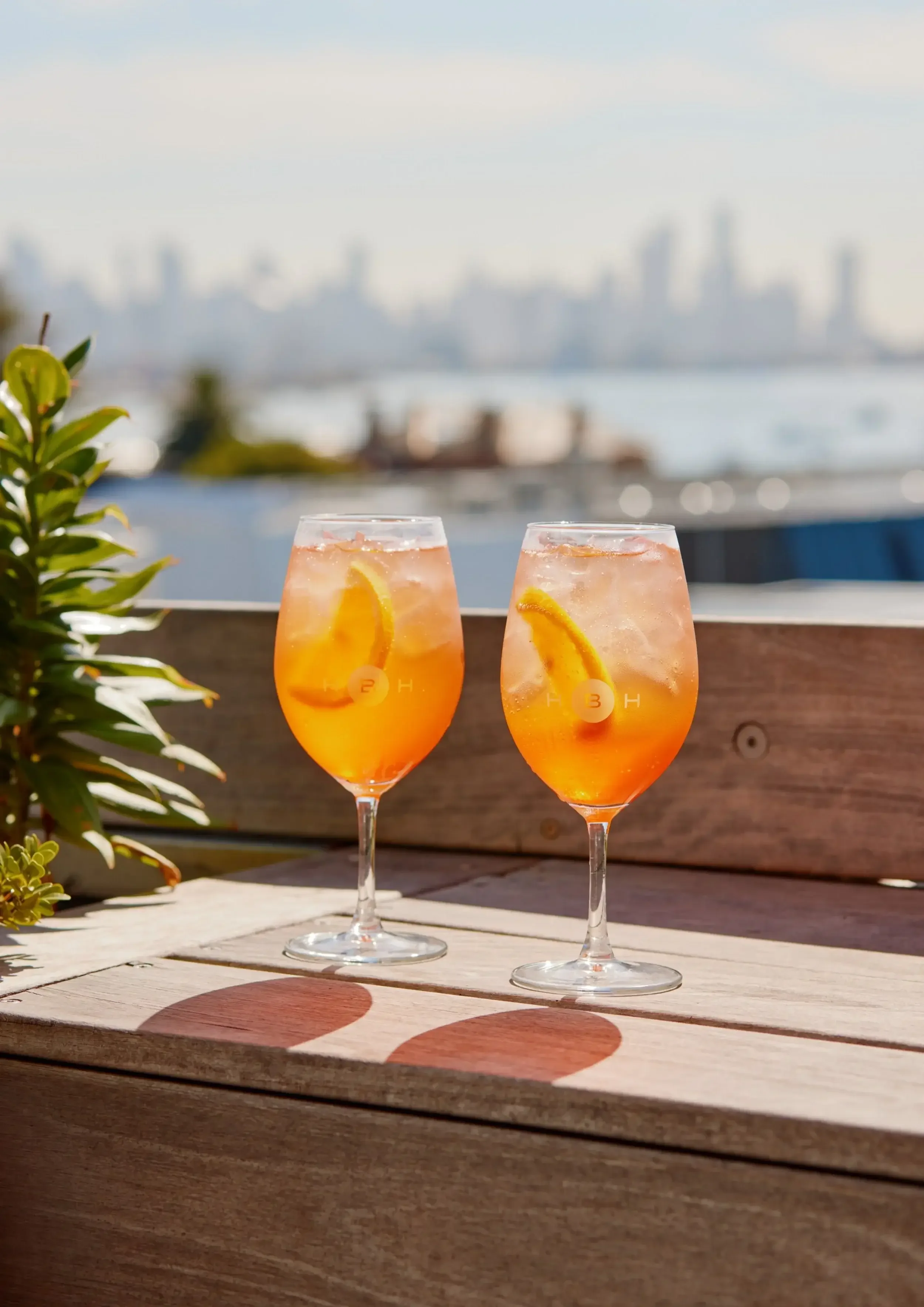 Two women enjoying drinks and toasting under a sun umbrella with a city skyline in the background.