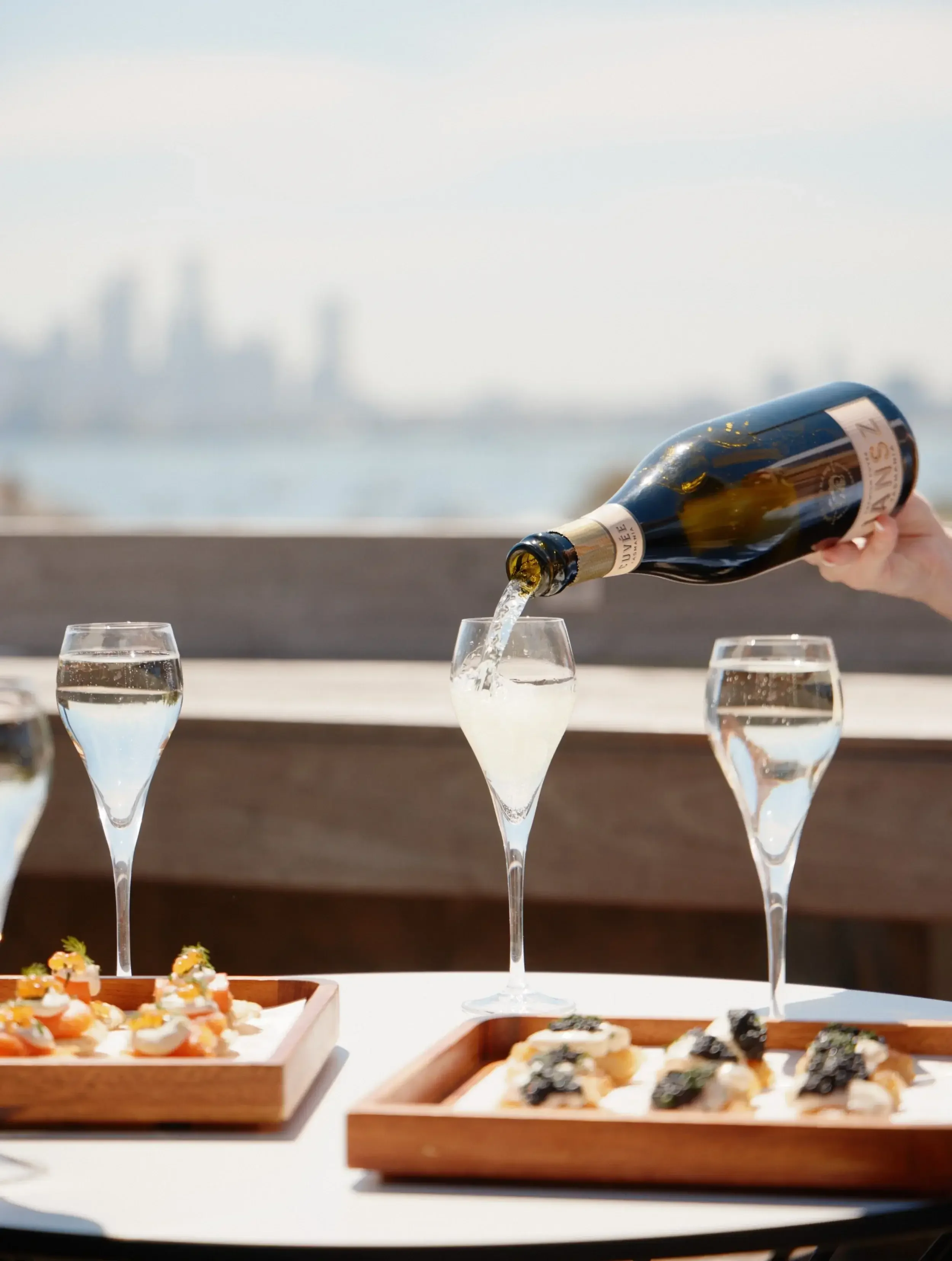 Person pouring champagne into a glass on an outdoor table with canapés and a city skyline in the background.