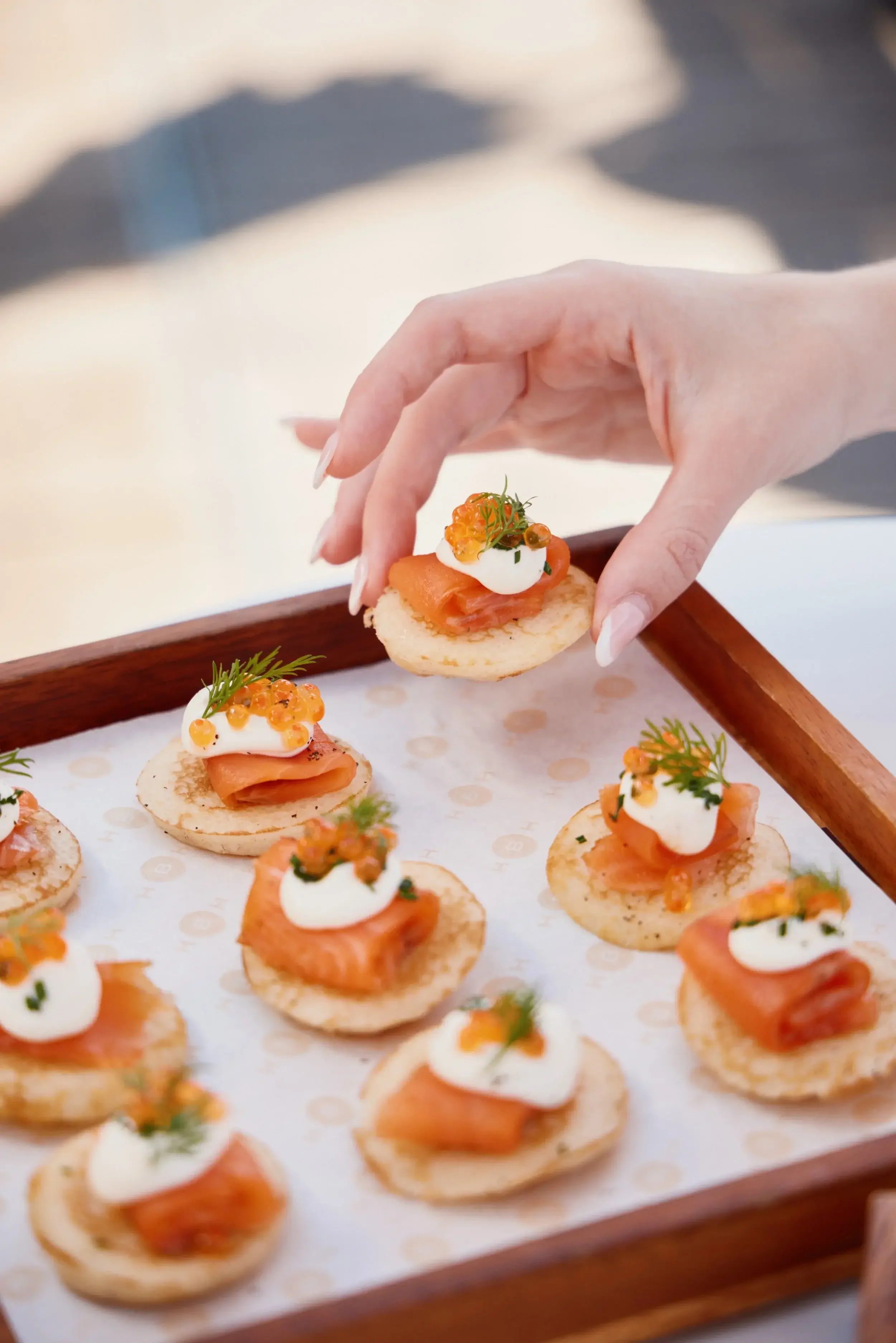 Hand picking up a smoked salmon canapé topped with cream, dill, and roe from a wooden serving tray.