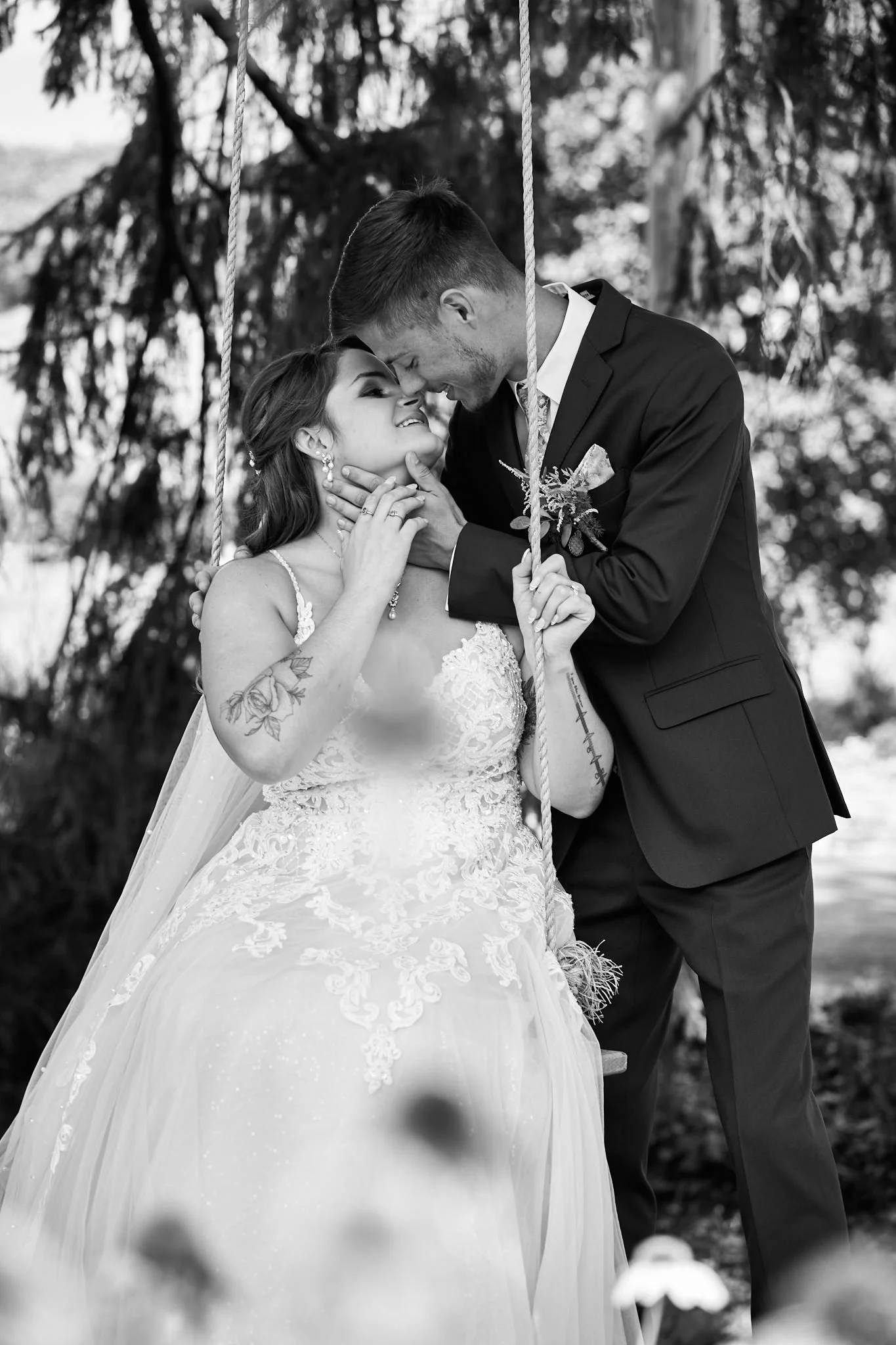 Black and white photo of a bride and groom sharing a close, intimate moment. The bride is seated on a swing, wearing a lace wedding dress, with tattoos visible on her arms. The groom is standing close, wearing a suit, gently holding the bride's face