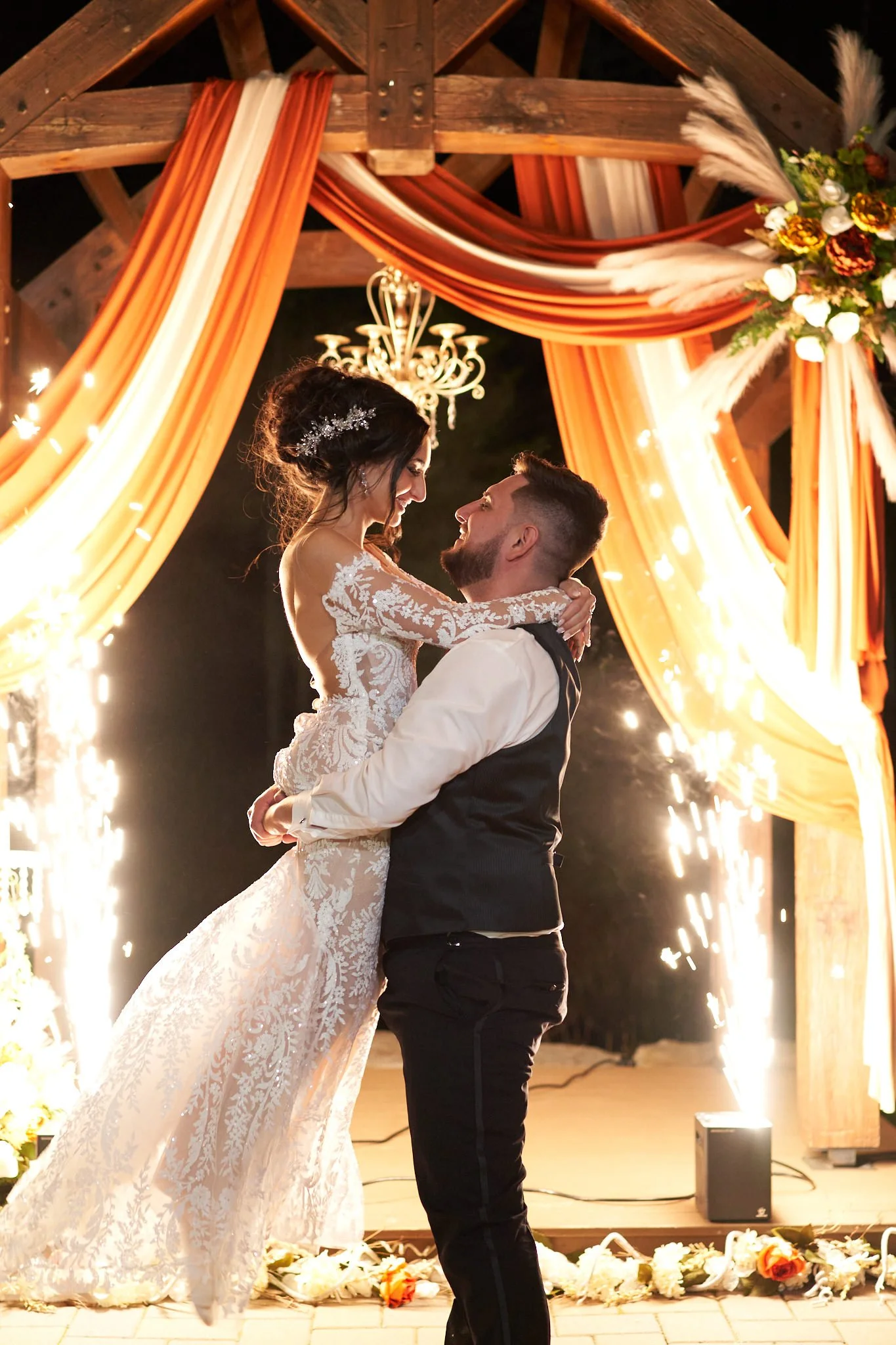 A bride and groom share a dance at their wedding under a decorated wooden arch with orange and beige drapes, flowers, and a chandelier, with sparklers burning in the background.