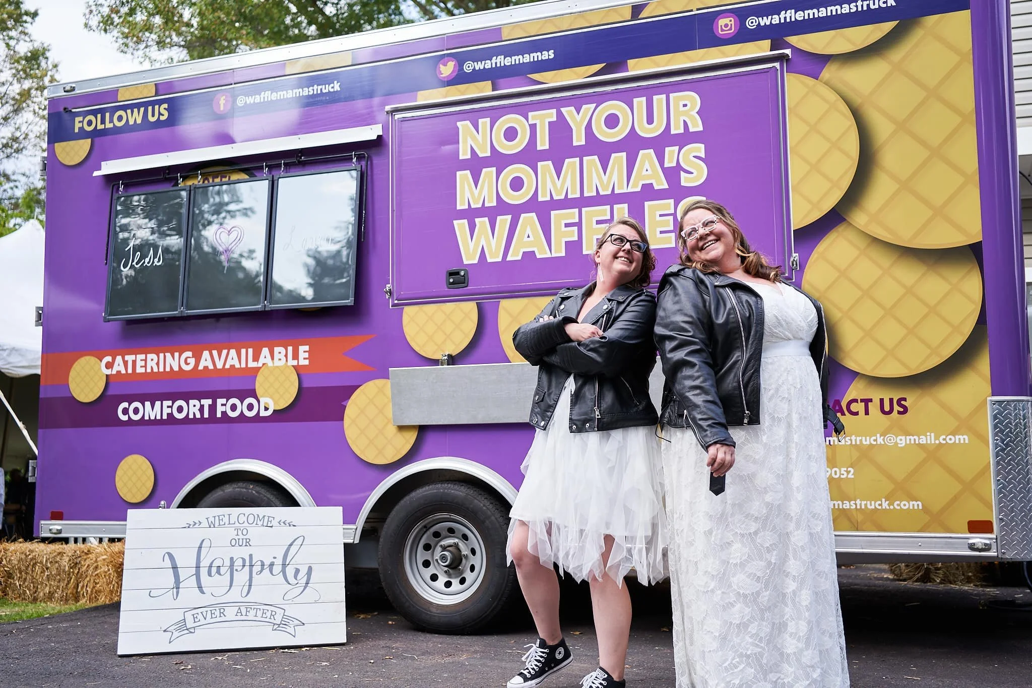 Two women in white dresses and black leather jackets standing in front of a purple waffle truck, smiling. The truck has signs indicating catering and comfort food, with the slogan 'Not your momma's waffle.' There is a welcome sign on the ground and h