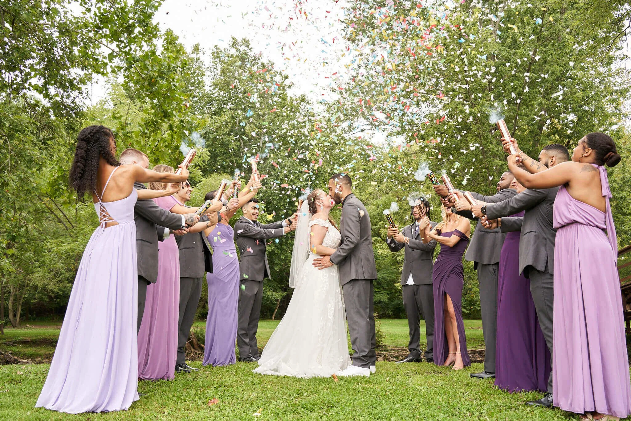 A wedding couple kissing surrounded by guests dressed in purple and gray, celebrating with confetti and party poppers outdoors.