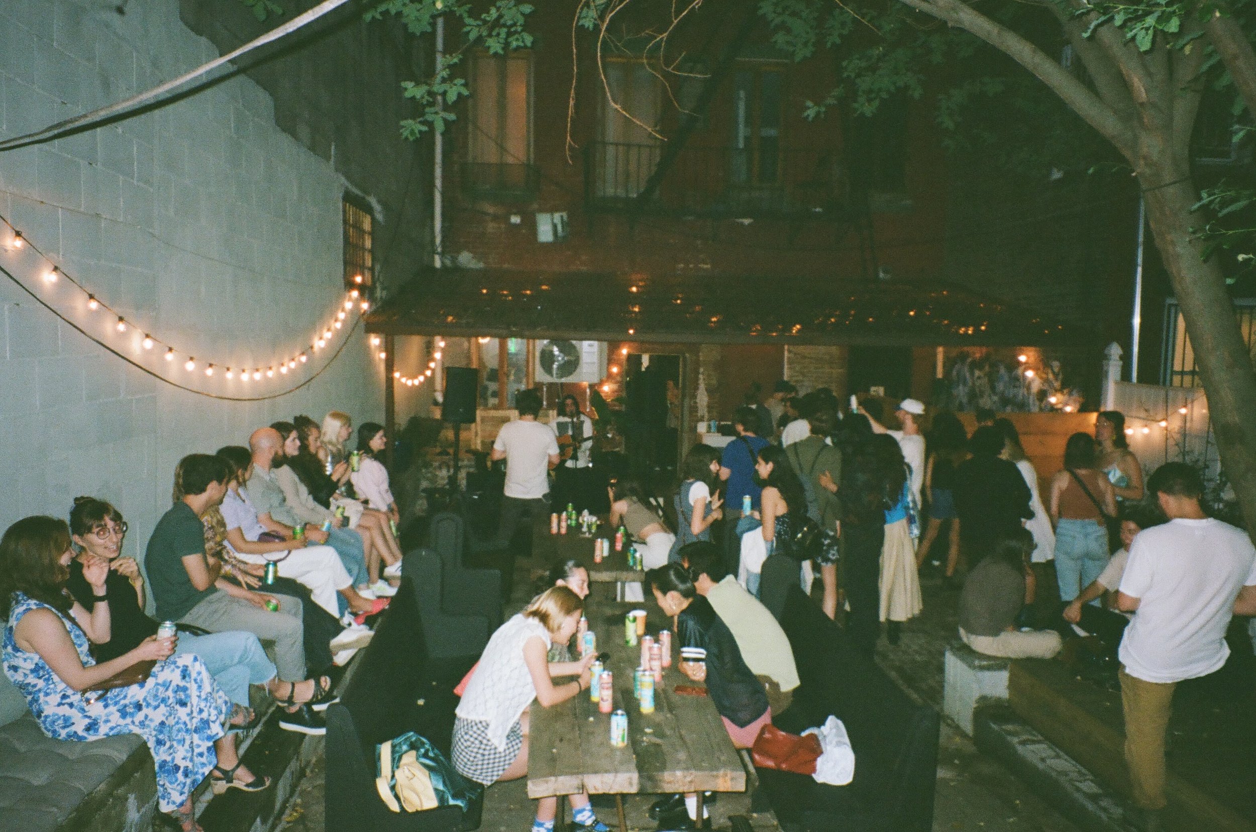 A large group of people gathered at a social event in an outdoor space, sitting and standing under string lights with trees and a brick wall in the background, during nighttime.