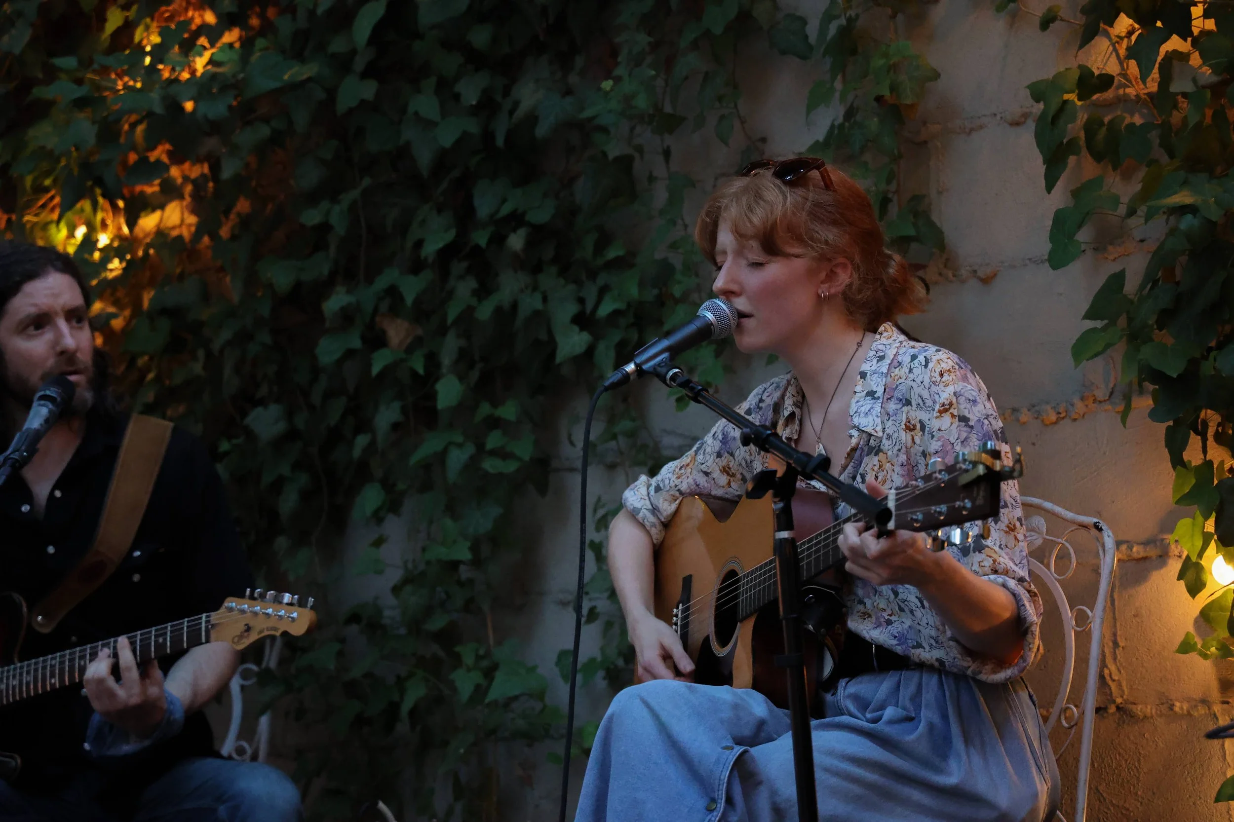 A woman with red hair singing and playing guitar with a microphone, a man with dark hair and beard playing guitar next to her, both seated outdoors against a green ivy-covered wall during the evening.
