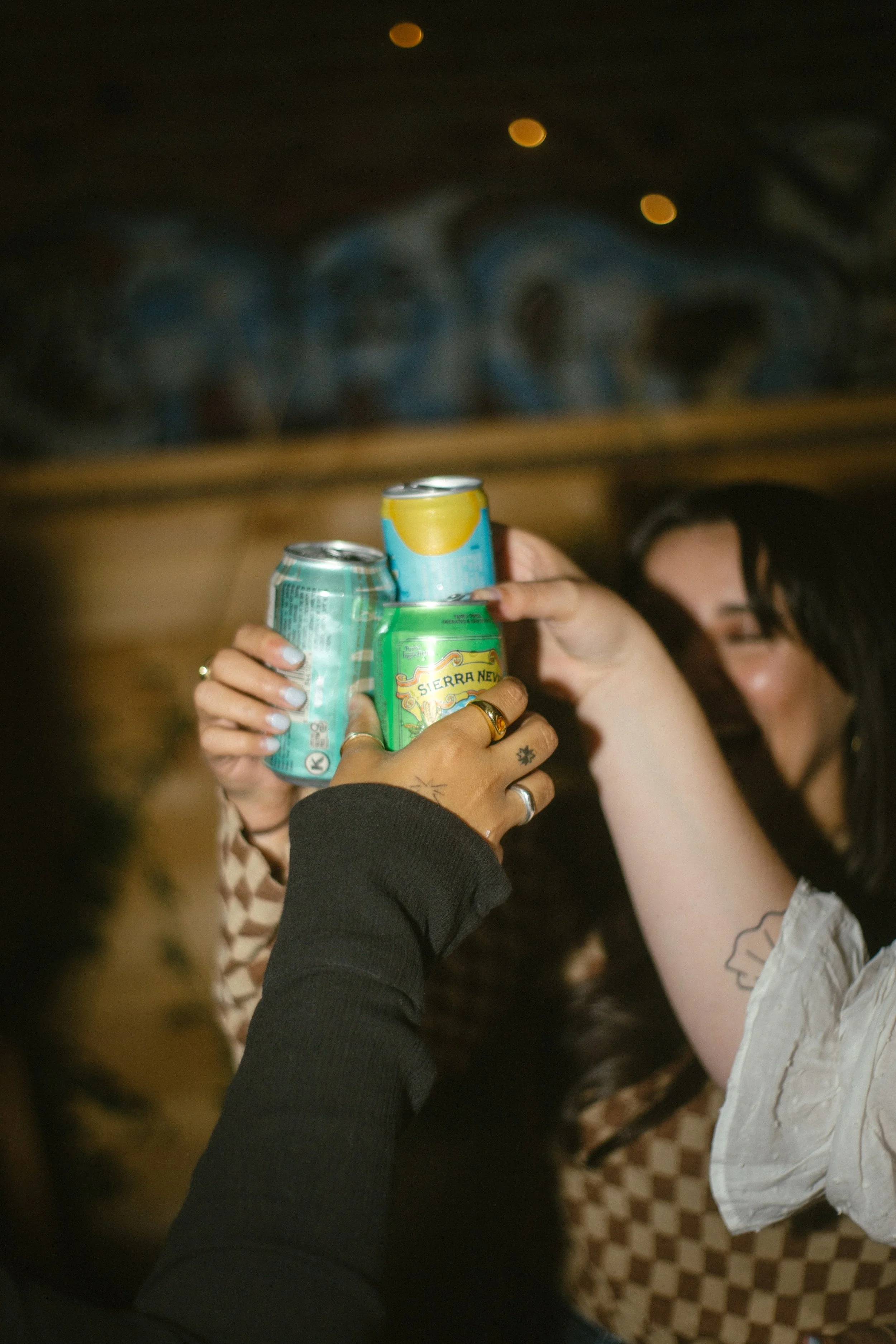 Multiple hands holding up cans of soda in a toast during a social gathering, with a woman smiling in the background.