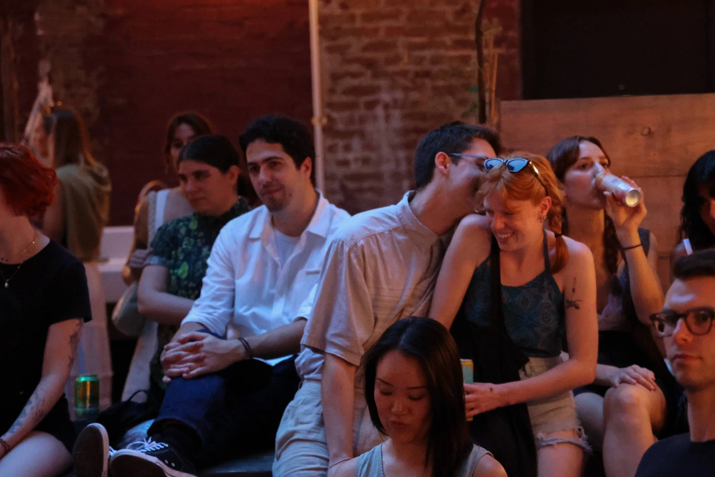 Group of people at a social gathering or party, sitting and standing in a dimly lit indoor space with brick walls, some holding drinks, smiling and talking.