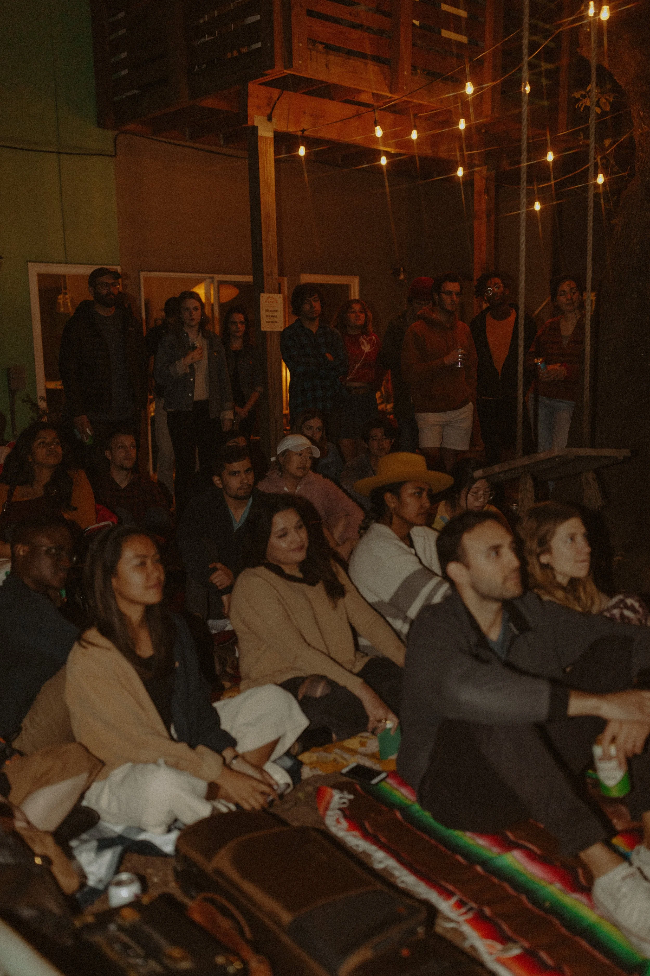 People gathered in an outdoor space at night, some standing and others seated, under string lights, with a wooden deck and green wall in the background.