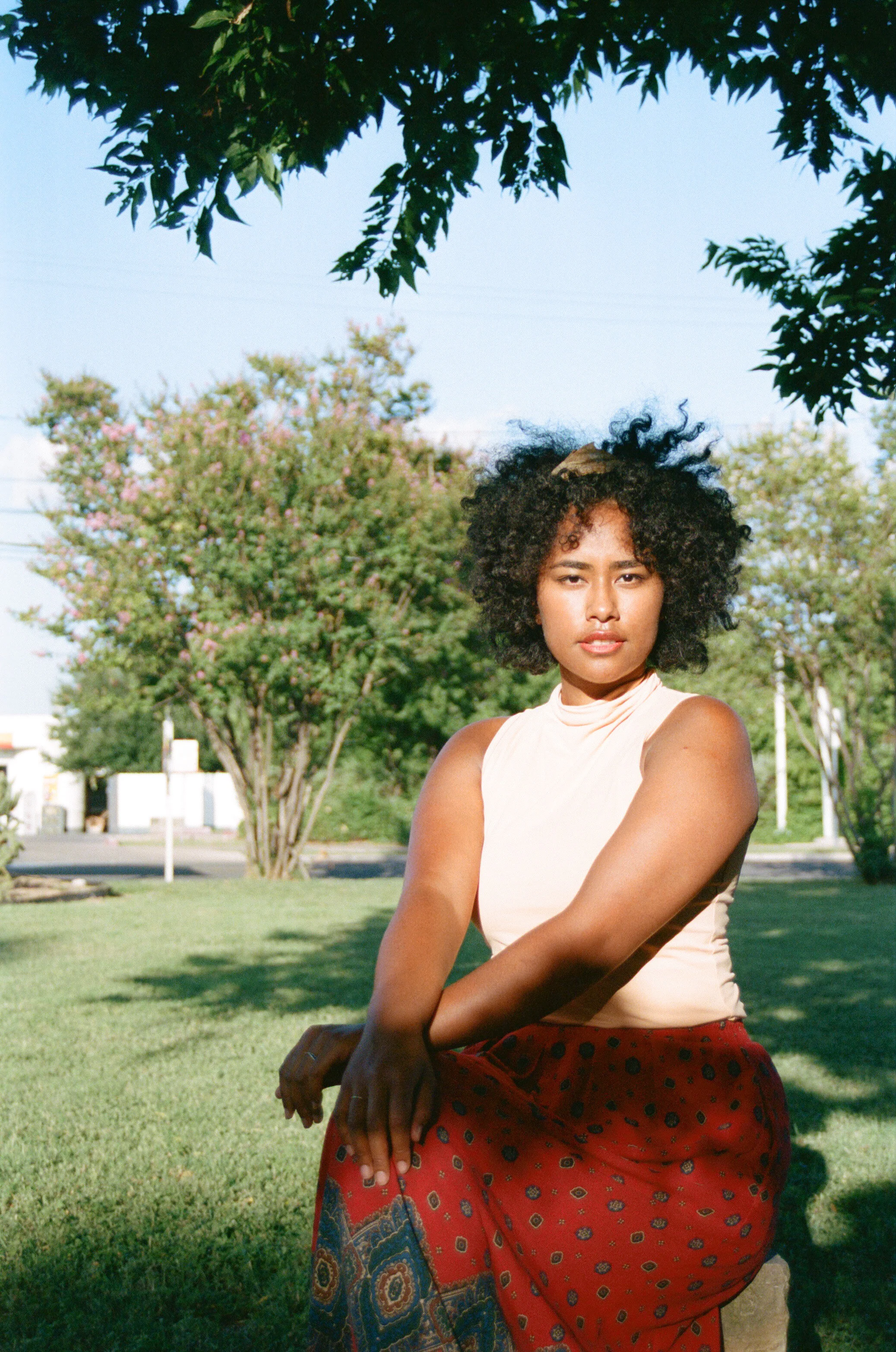 A woman with curly hair and a light sleeveless top sitting outdoors in a park-like setting with trees, greenery, and flowering trees in the background.