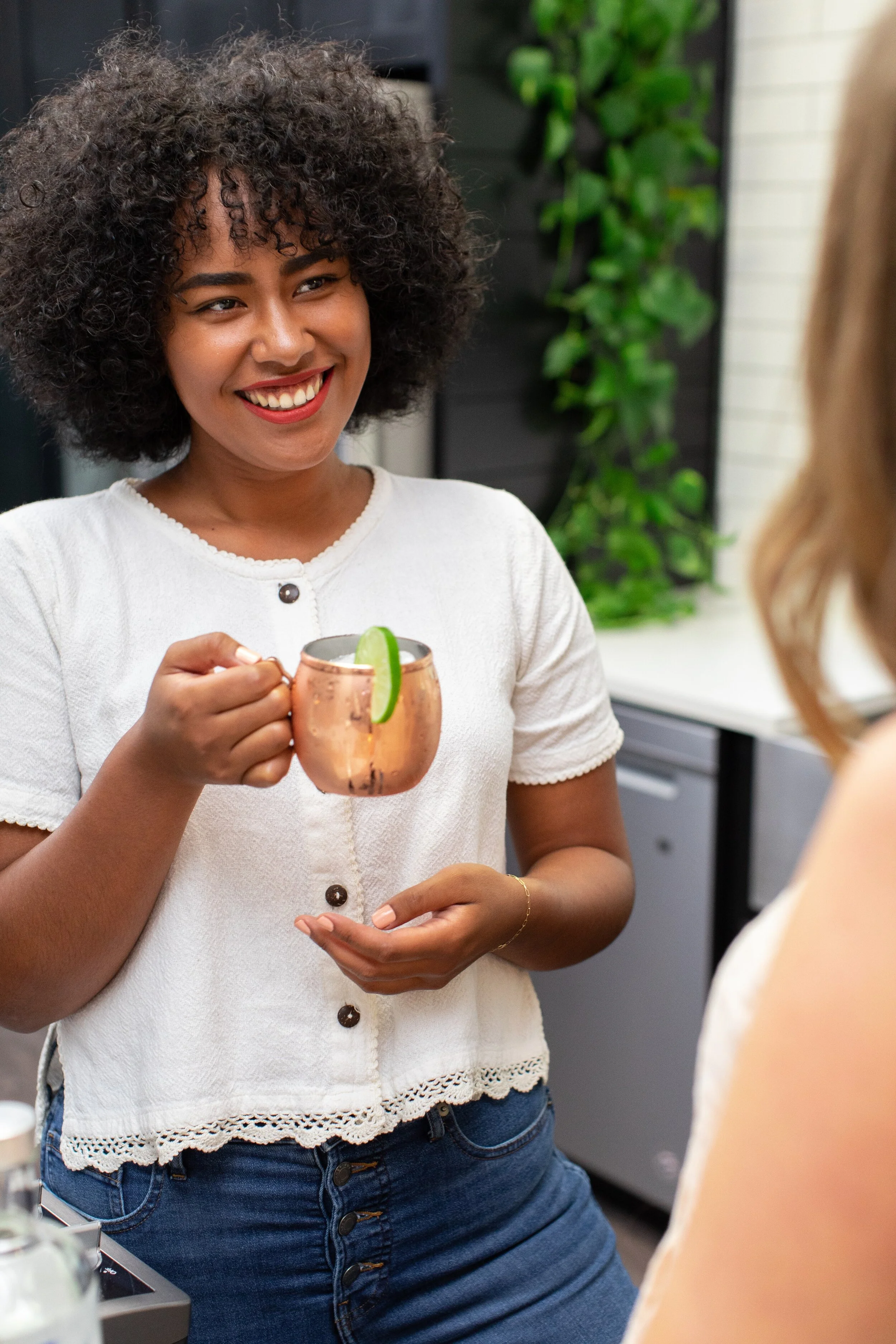 A woman with curly hair, smiling, holding a copper mug with a lime wedge, engaging in conversation with someone outside the frame.