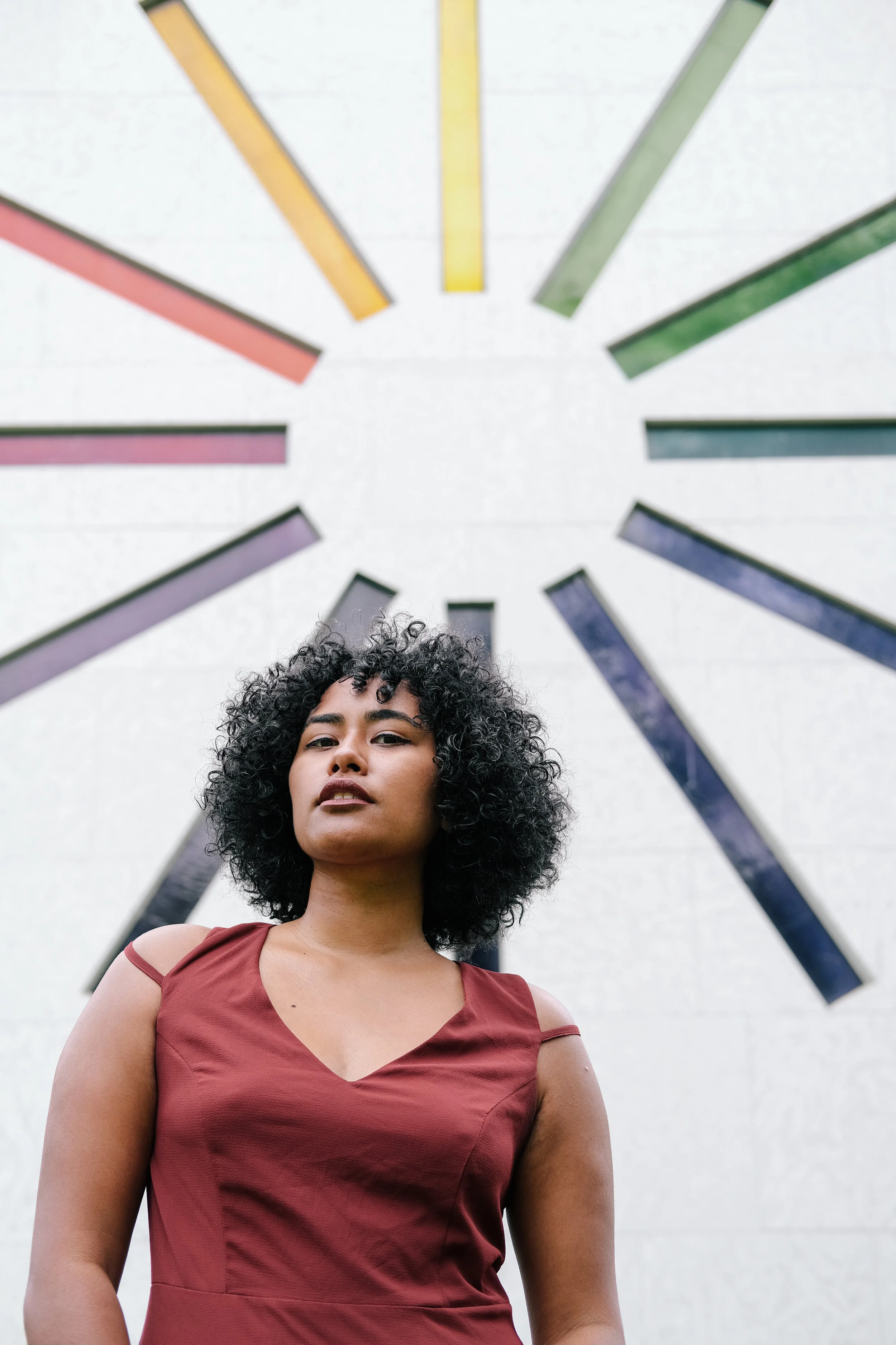 A woman with curly black hair and tan skin wearing a sleeveless maroon dress stands outdoors in front of a white wall with colorful, elongated geometric shapes arranged in a circular pattern above her head.