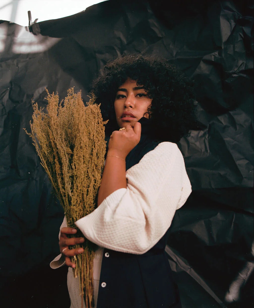 A woman with curly black hair lying on a black background, holding a bouquet of dried tan-colored flowers.