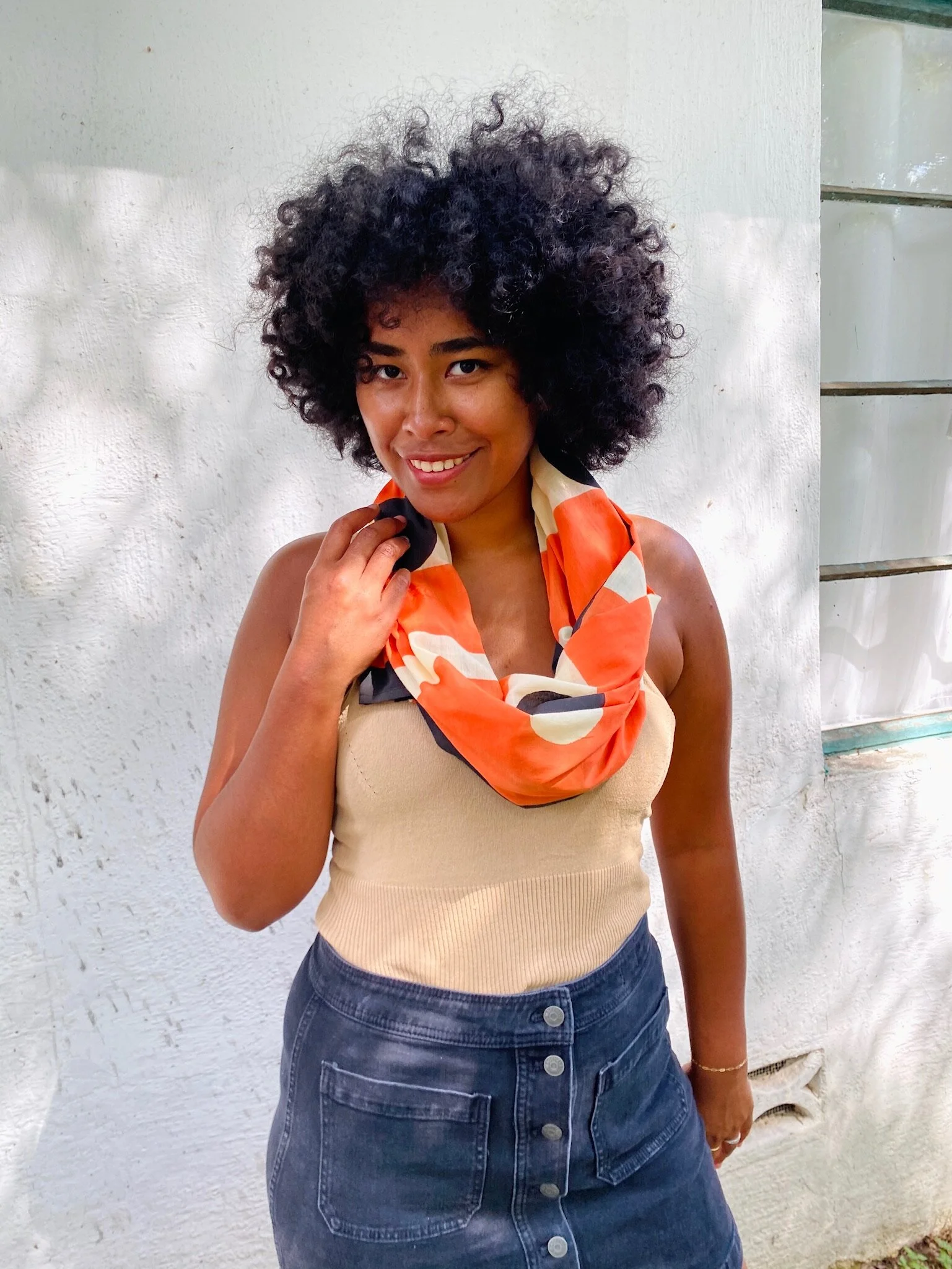 A woman with dark curly hair, wearing a beige tank top and a colorful scarf around her neck, standing in front of a white wall, smiling and holding the scarf with her right hand.