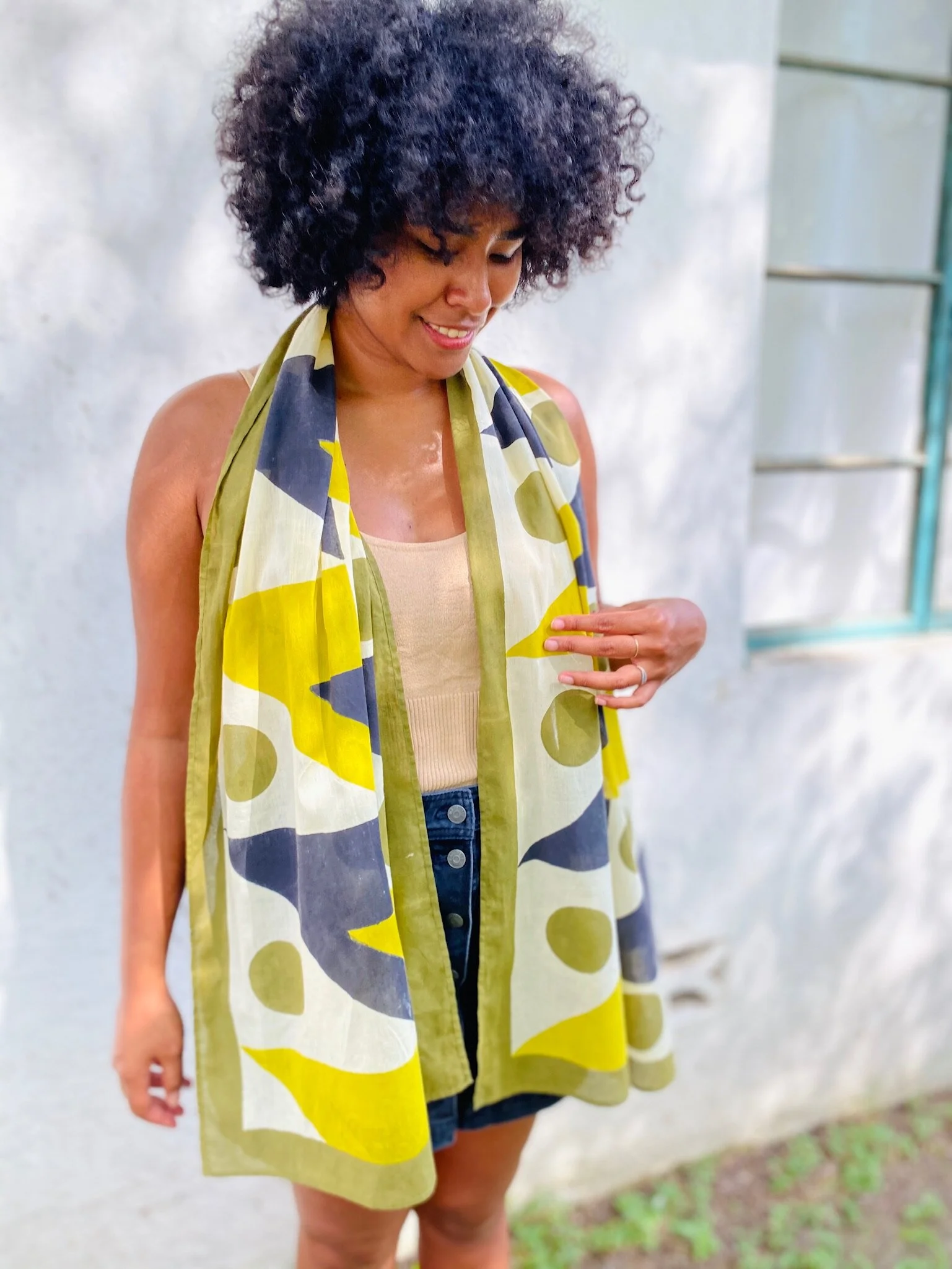 A woman with curly black hair wearing a beige tank top and a multicolored scarf stands outdoors near a white wall, smiling and looking down.