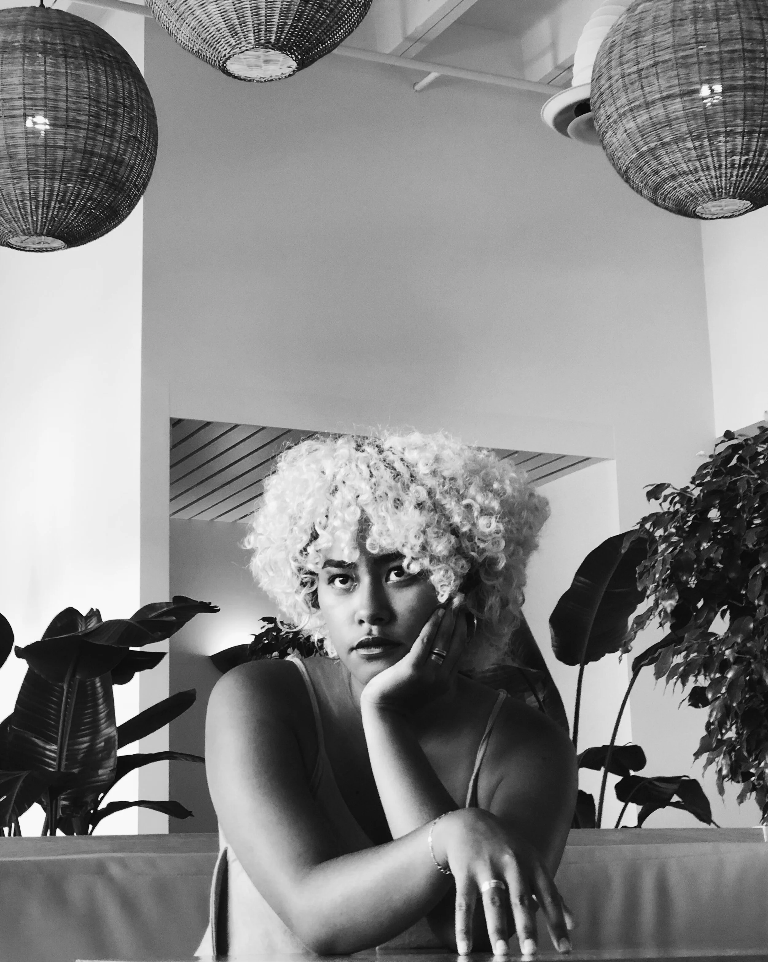 Black and white photo of a woman with curly hair sitting at a table, with plants around her, resting her head on her hand, and looking directly at the camera.