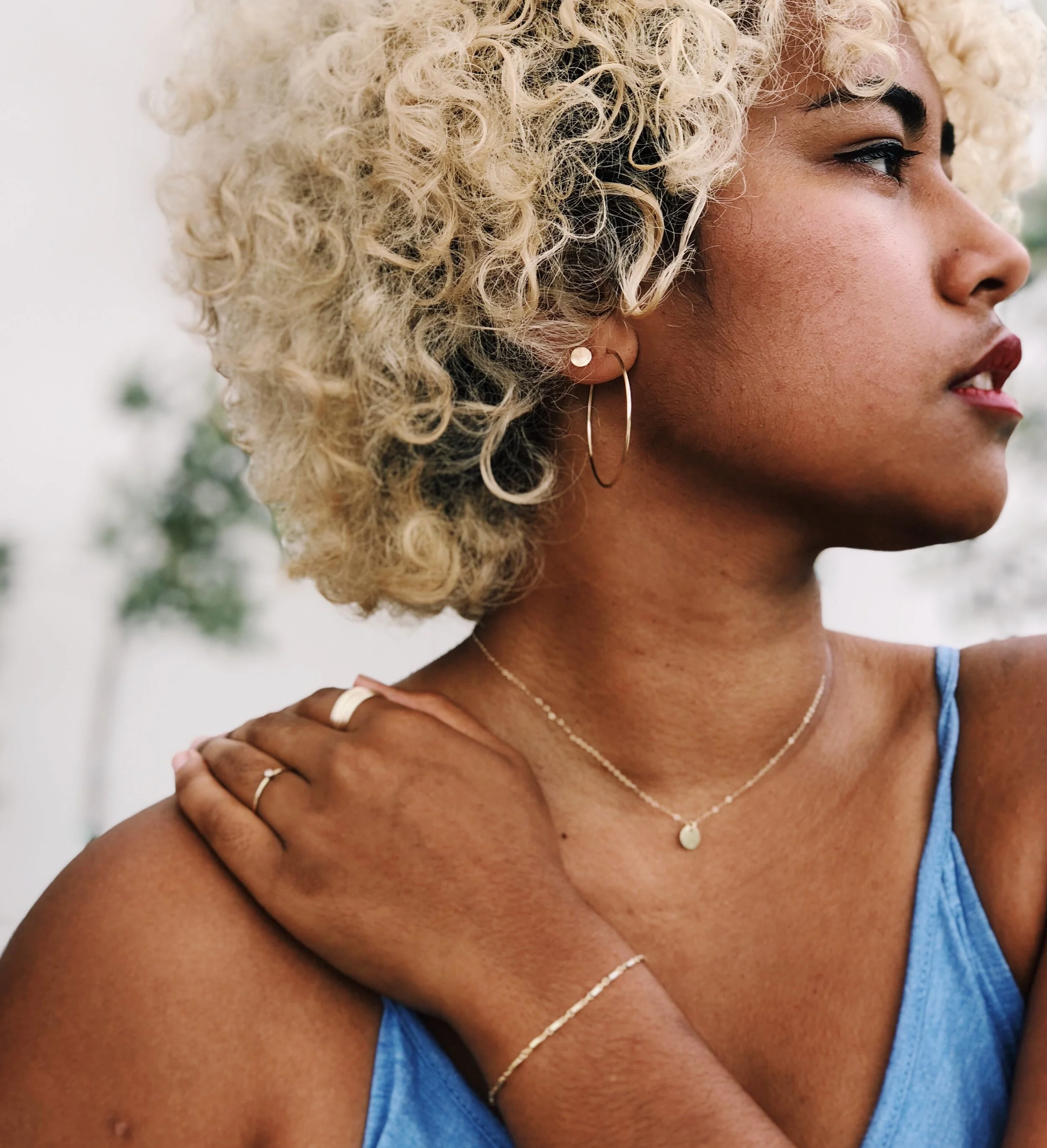 A woman with curly blonde hair, tan skin, wearing jewelry, a blue top, and looking to the side.