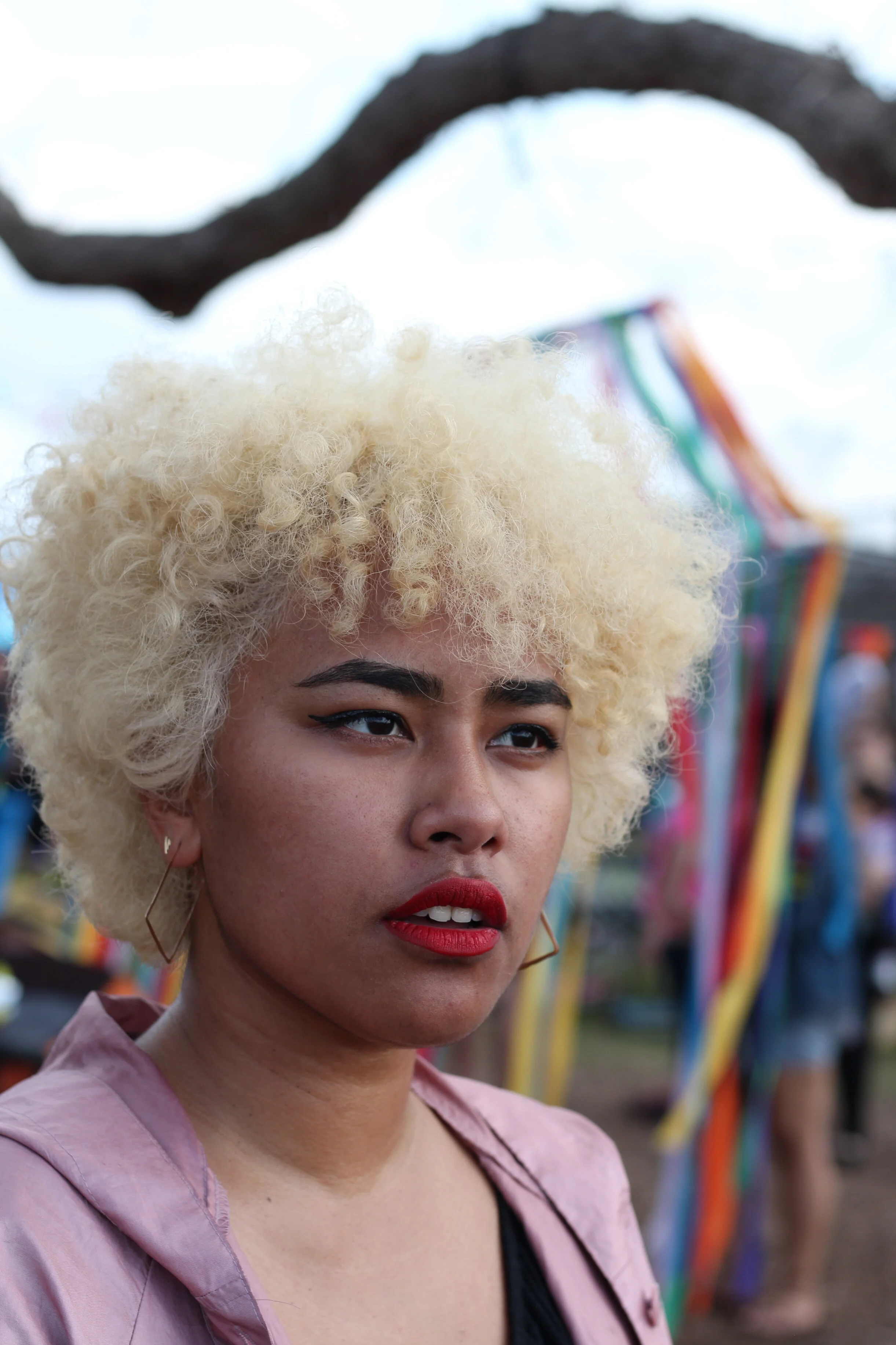 A woman with platinum blonde, curly hair and bold red lipstick, wearing a pink jacket and black top, looking seriously at the camera with a blurred outdoor festival background.