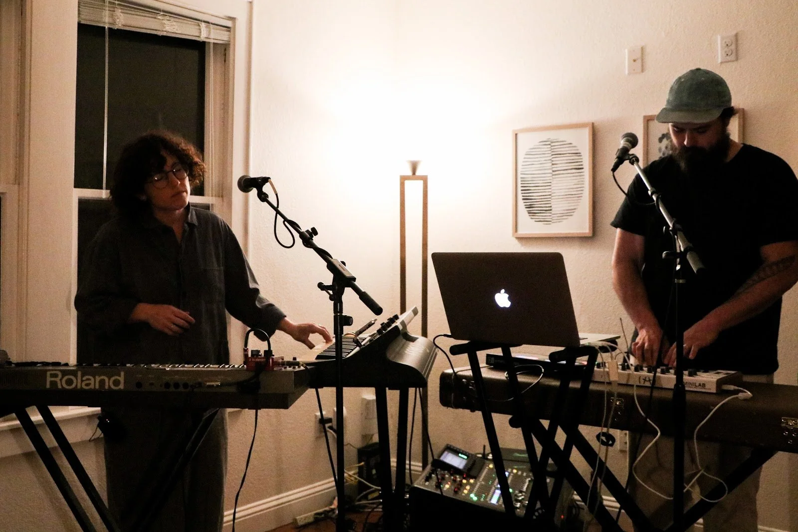 Two musicians performing with electronic equipment in a cozy indoor setting, one woman using a keyboard and the other man adjusting sound gear, with artwork on the wall and warm lighting.