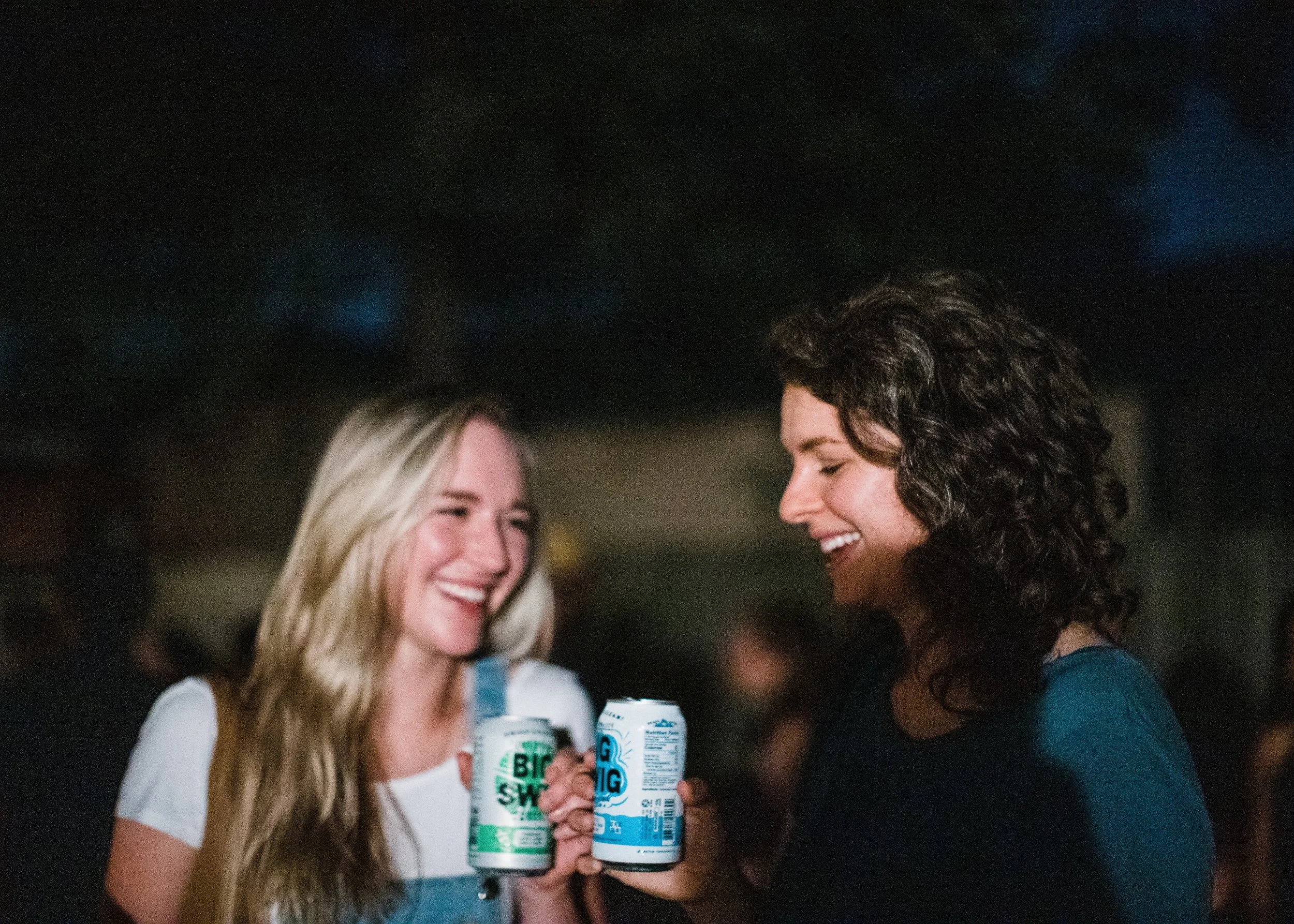 Two women smiling and holding cans of a beverage together at a social gathering or party.