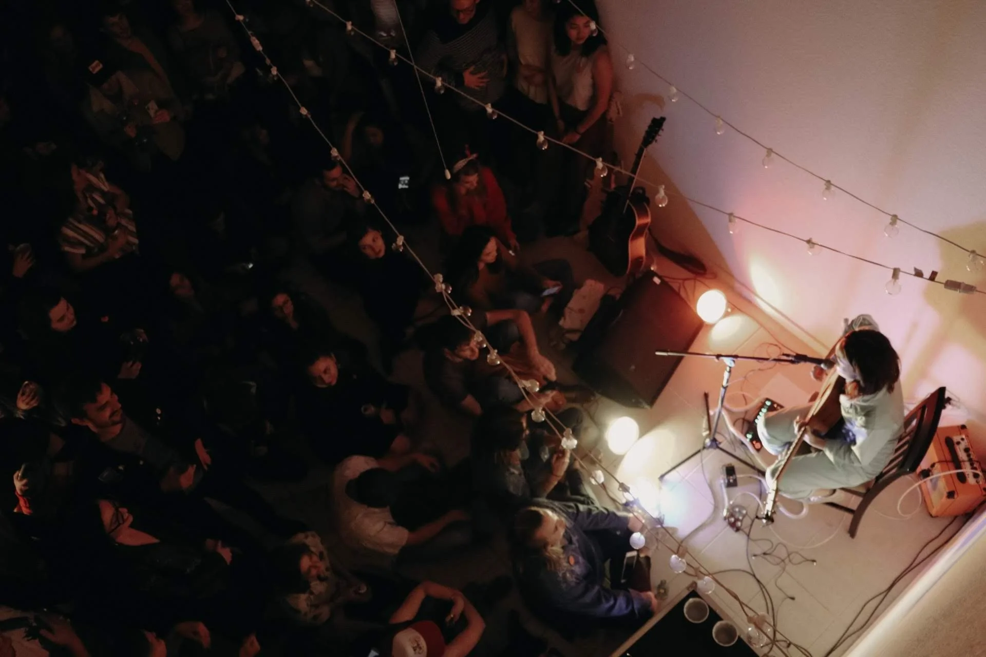 An indoor live music performance where a musician with long hair, dressed in white, is playing an acoustic guitar on a small stage. Audience members are seated and standing, facing the musician, with string lights hanging overhead creating a cozy atm