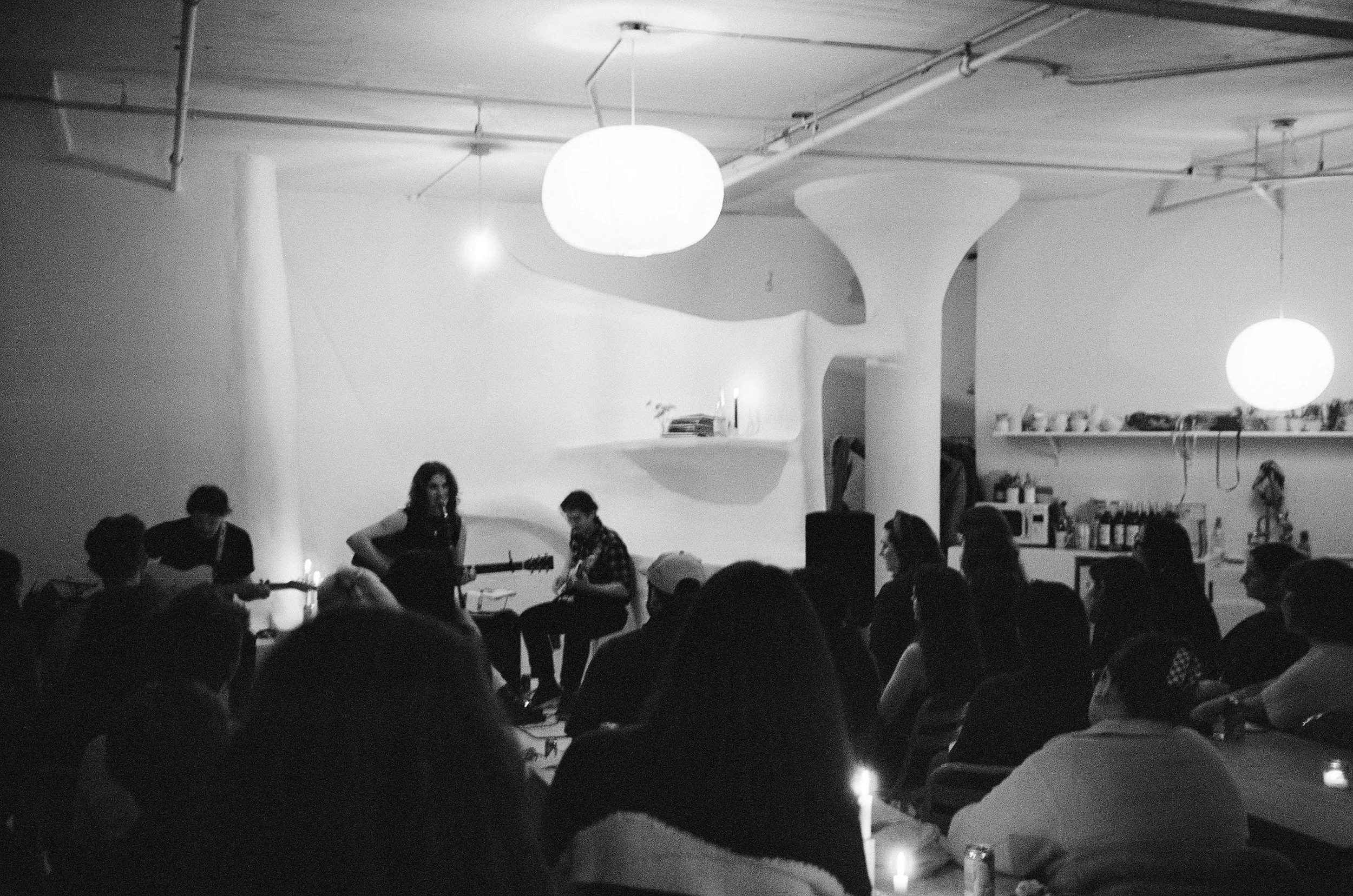 A black and white photo of an indoor musical performance with a group of people. There are hanging spherical lights from the ceiling, a large abstract wall sculpture, and books or objects on shelves.