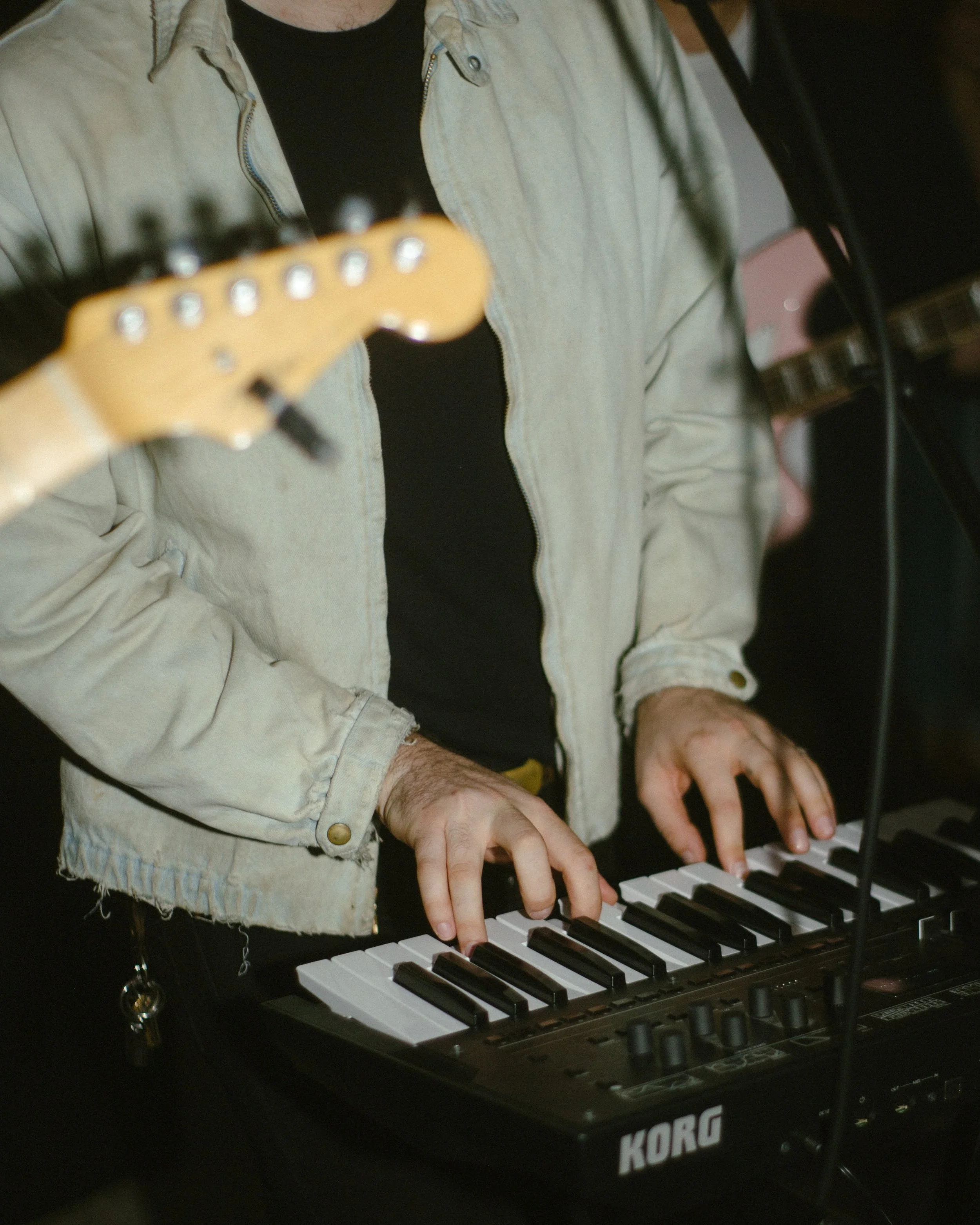 Person playing a keyboard with their hands, wearing a beige jacket over a black shirt, and standing in front of a Korg keyboard synthesizer.