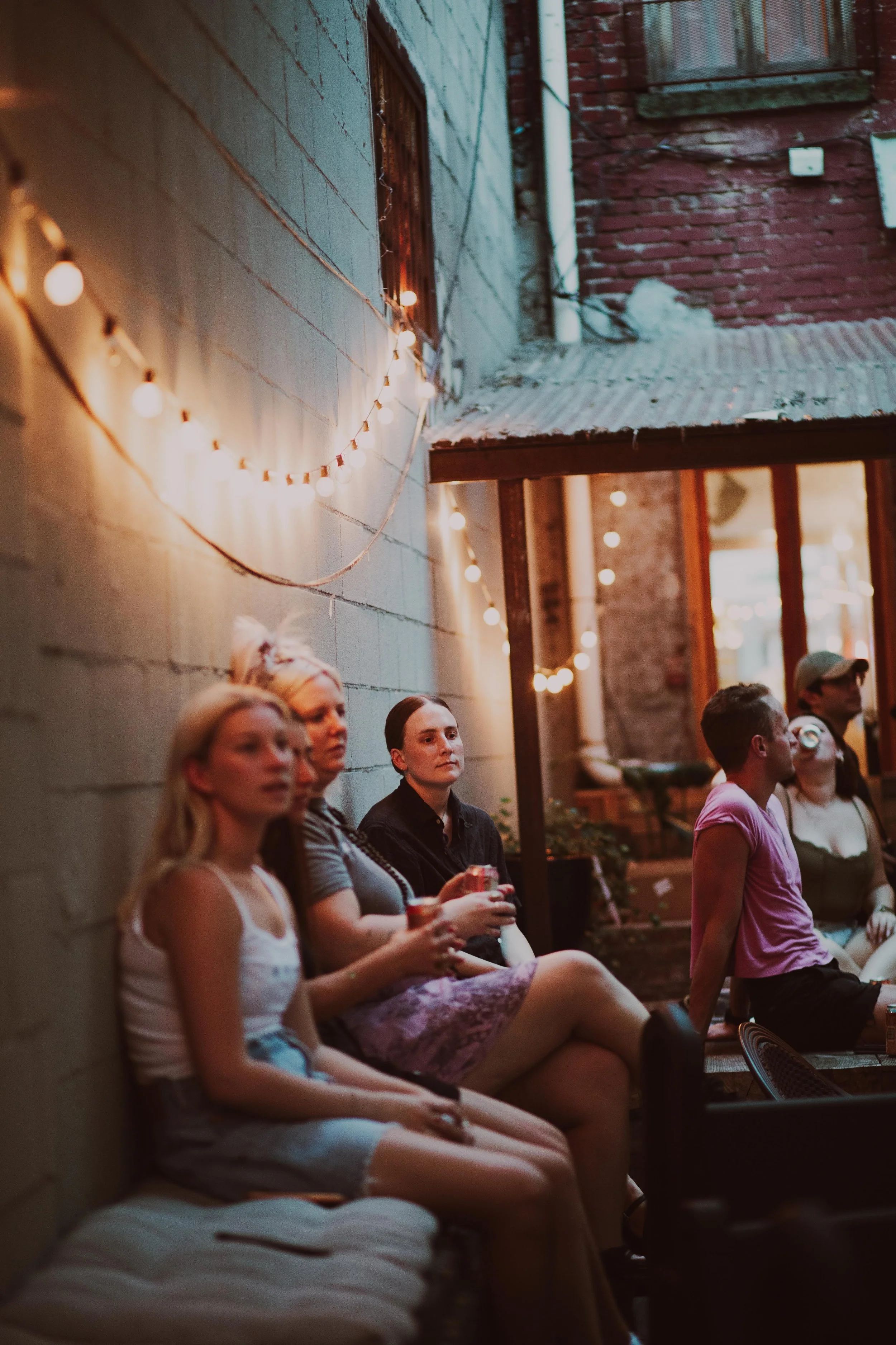 Group of people sitting outdoors in a semi-covered area with string lights, brick wall, and windows in the background.