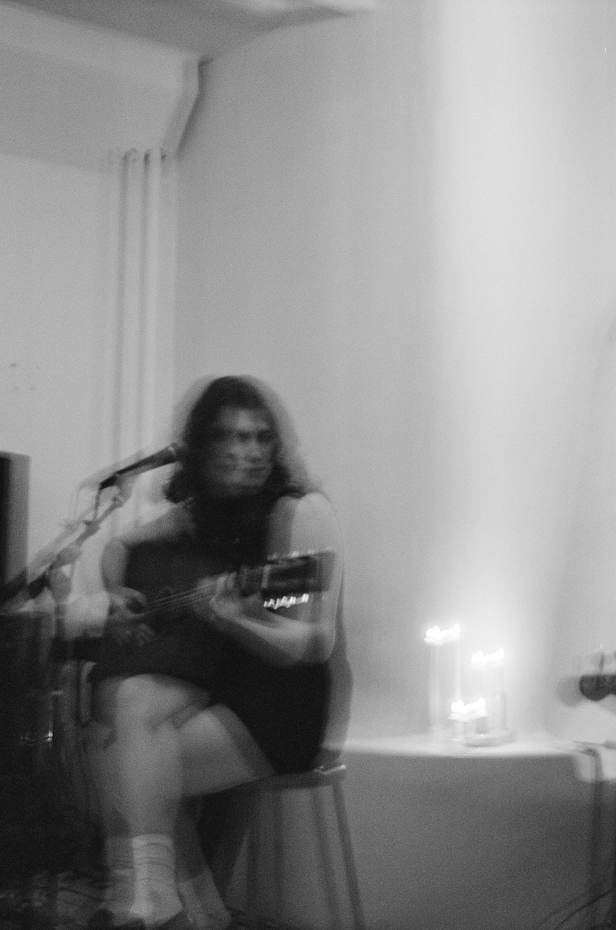 A woman with curly hair and sunglasses is sitting on a chair playing an acoustic guitar. There are lit candles on a table to her right, and the wall behind her is plain with a curtained window.
