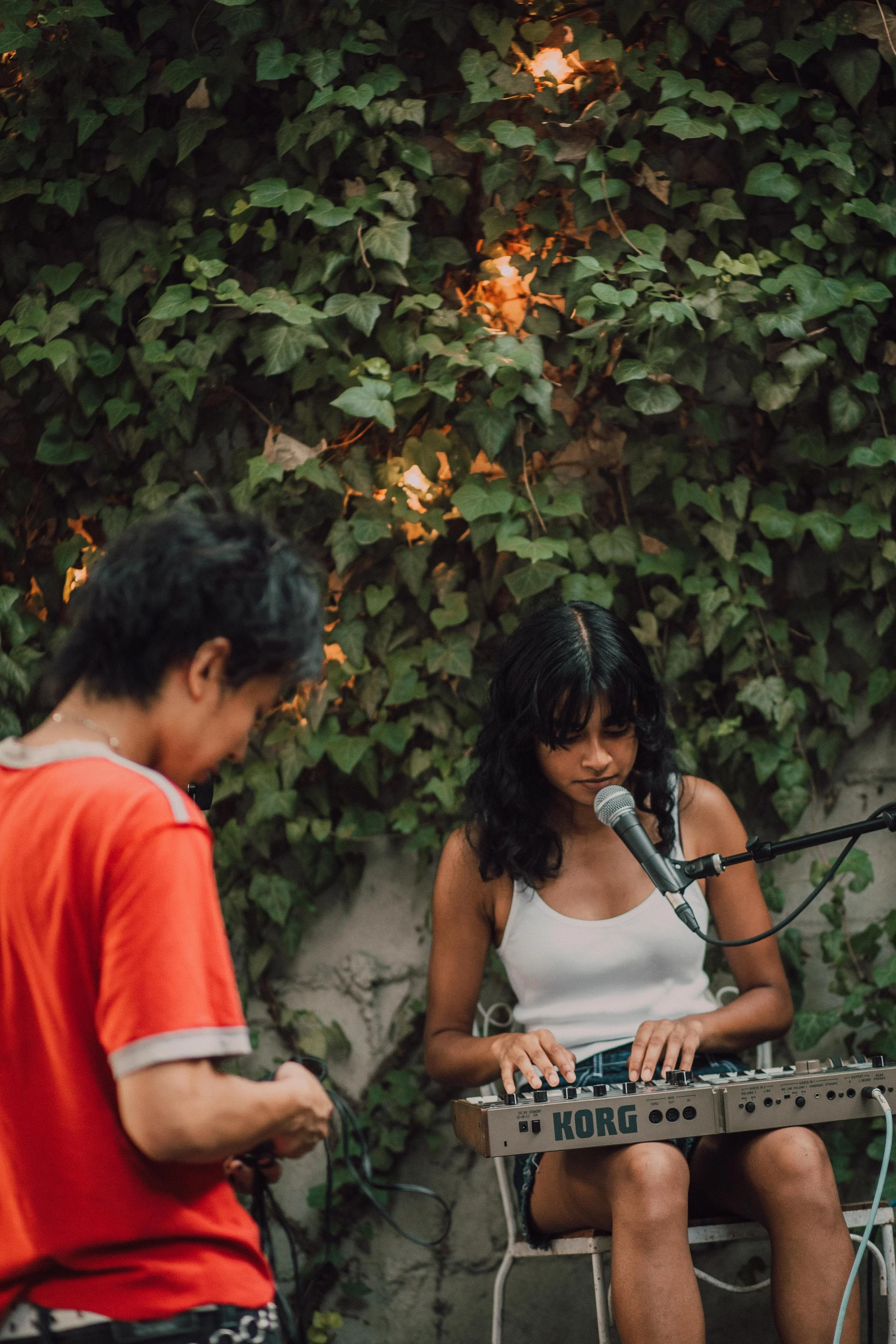 A woman with black hair playing a keyboard, sitting outdoors against a leafy green wall, while a man in a red shirt stands nearby.