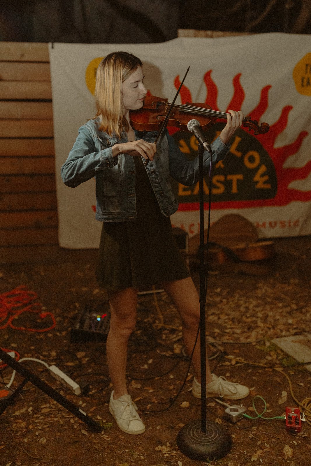 A young woman playing the violin on stage with a microphone in front of her, wearing a denim jacket, black dress, and sneakers, with a backdrop featuring red flames and black text.