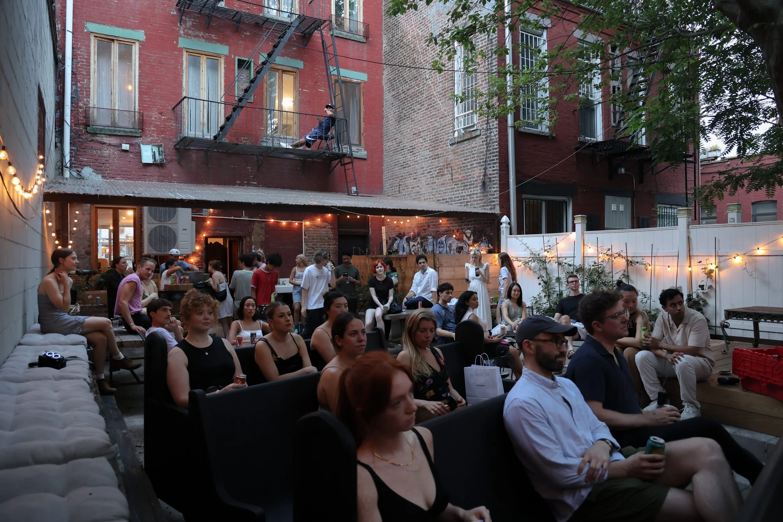 A gathering of people seated in an outdoor patio or courtyard with string lights, behind a white fence, in a city setting, with brick buildings and trees in the background, during evening.