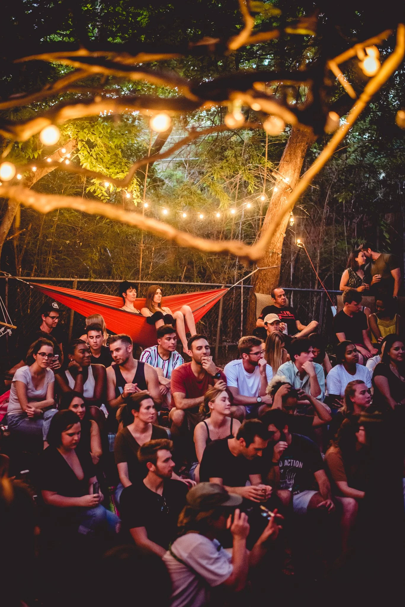 A group of people gathered outdoors at night, seated and standing, with trees and string lights overhead. Some are engaged in conversation, while others are looking towards the front.