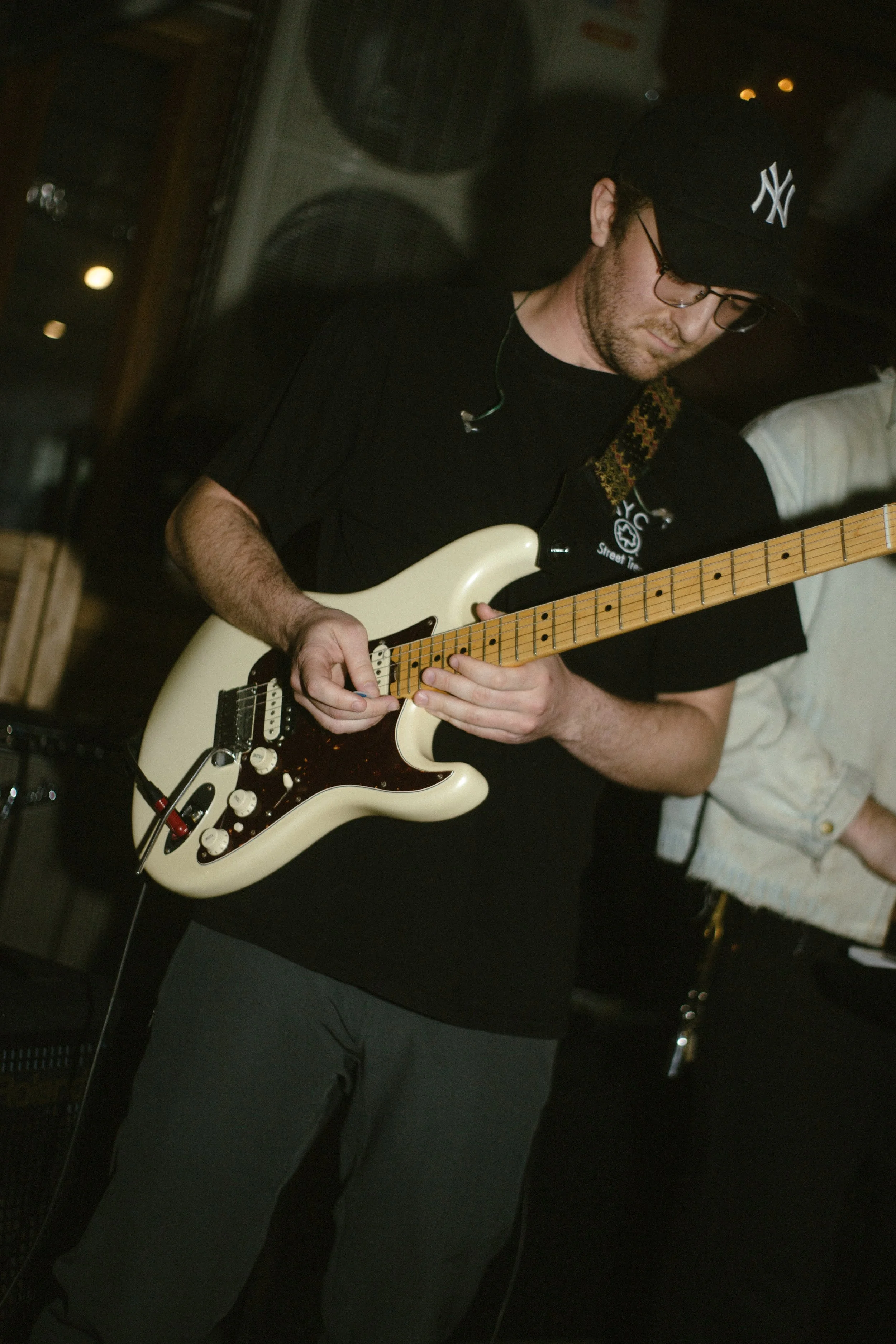A man wearing a black baseball cap with a white 'NY' logo, glasses, and a black T-shirt playing a cream-colored electric guitar in a dimly lit setting.