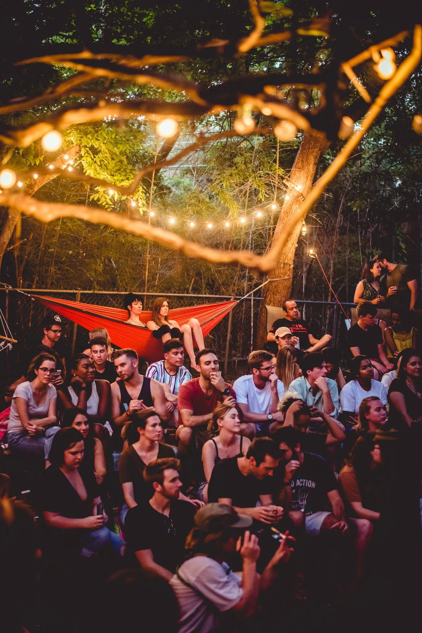 A large gathering of people seated outdoors at dusk, with string lights hanging from trees, some individuals are engaged in conversation while others are looking at their phones.